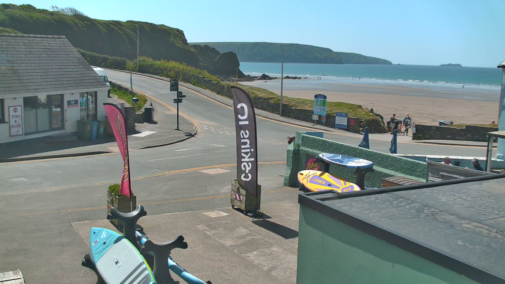 A sunny day at a beach shows a sandy shore with people, a calm ocean, and a road leading to a building near the water.