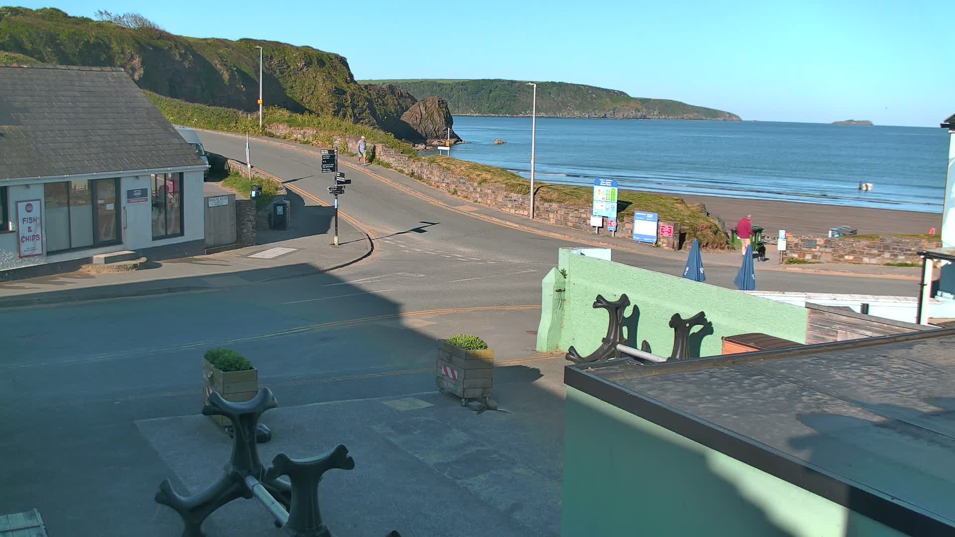 A sunny coastal scene shows a road leading down to a sandy beach with calm ocean waters and green hills in the background.