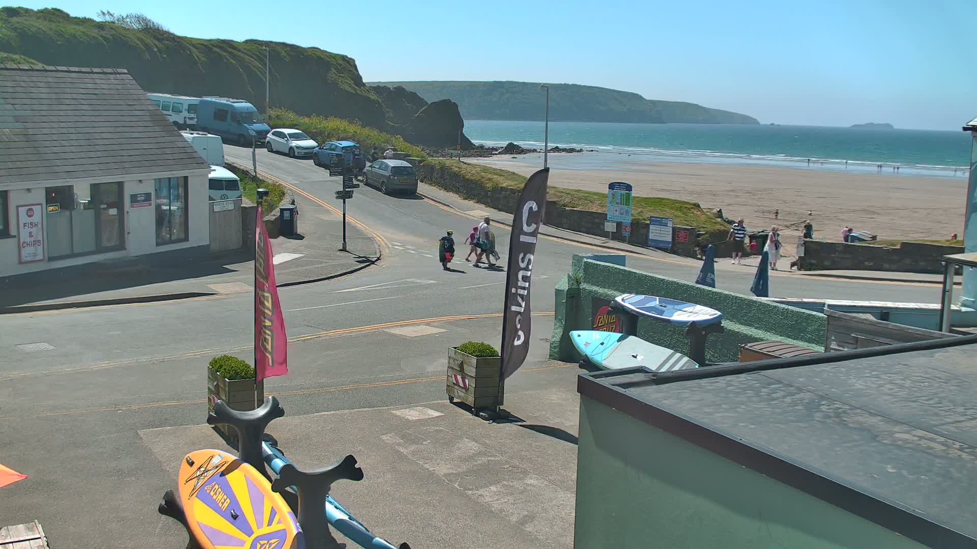 A sunny day at a beachside town shows a sandy beach with a few people on it, cars parked near buildings, and surfboards near a roadway.