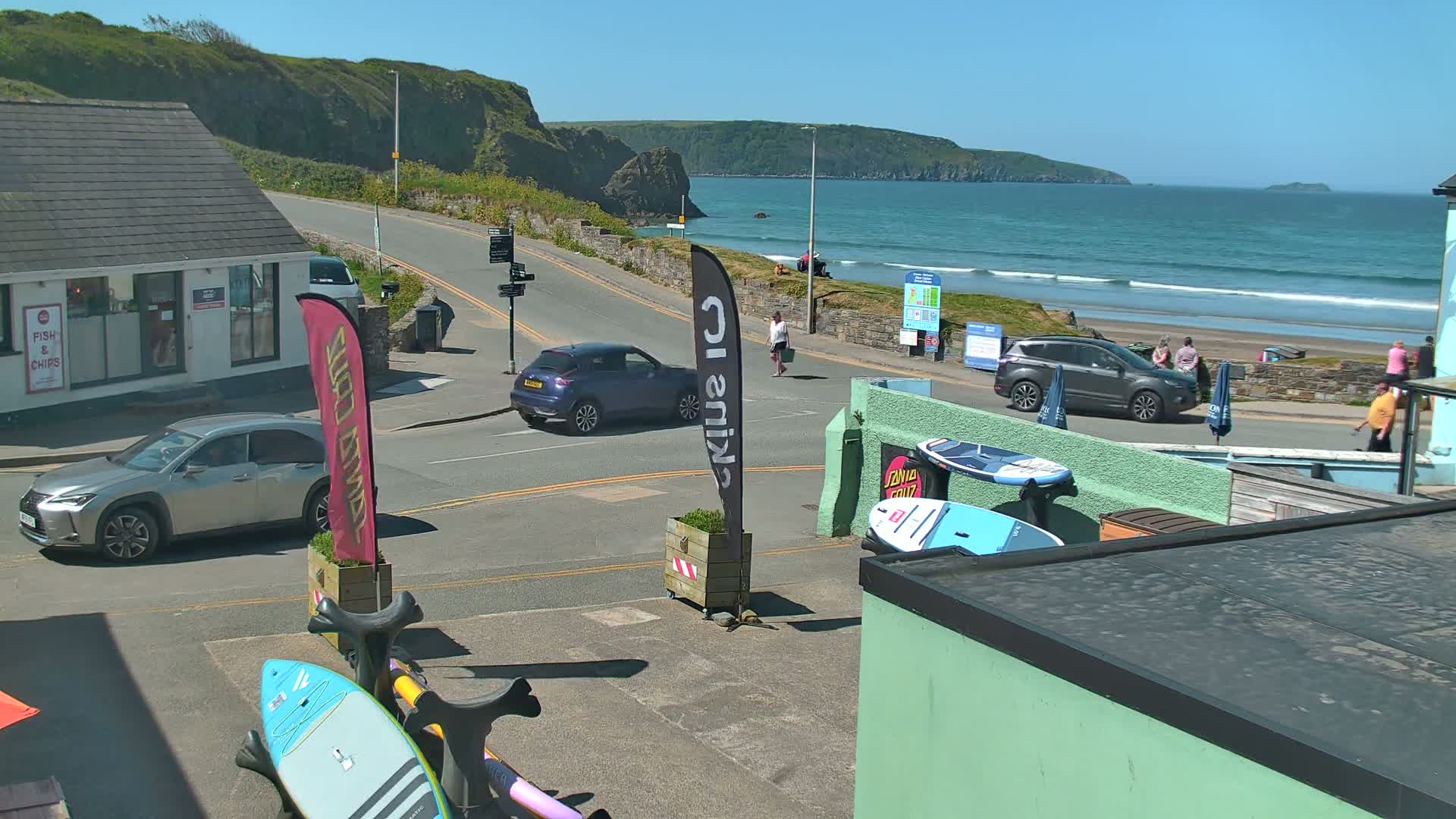 A sunny seaside town scene shows a road with cars, pedestrians, and several surfboards leaning against a building overlooking a beach with waves.