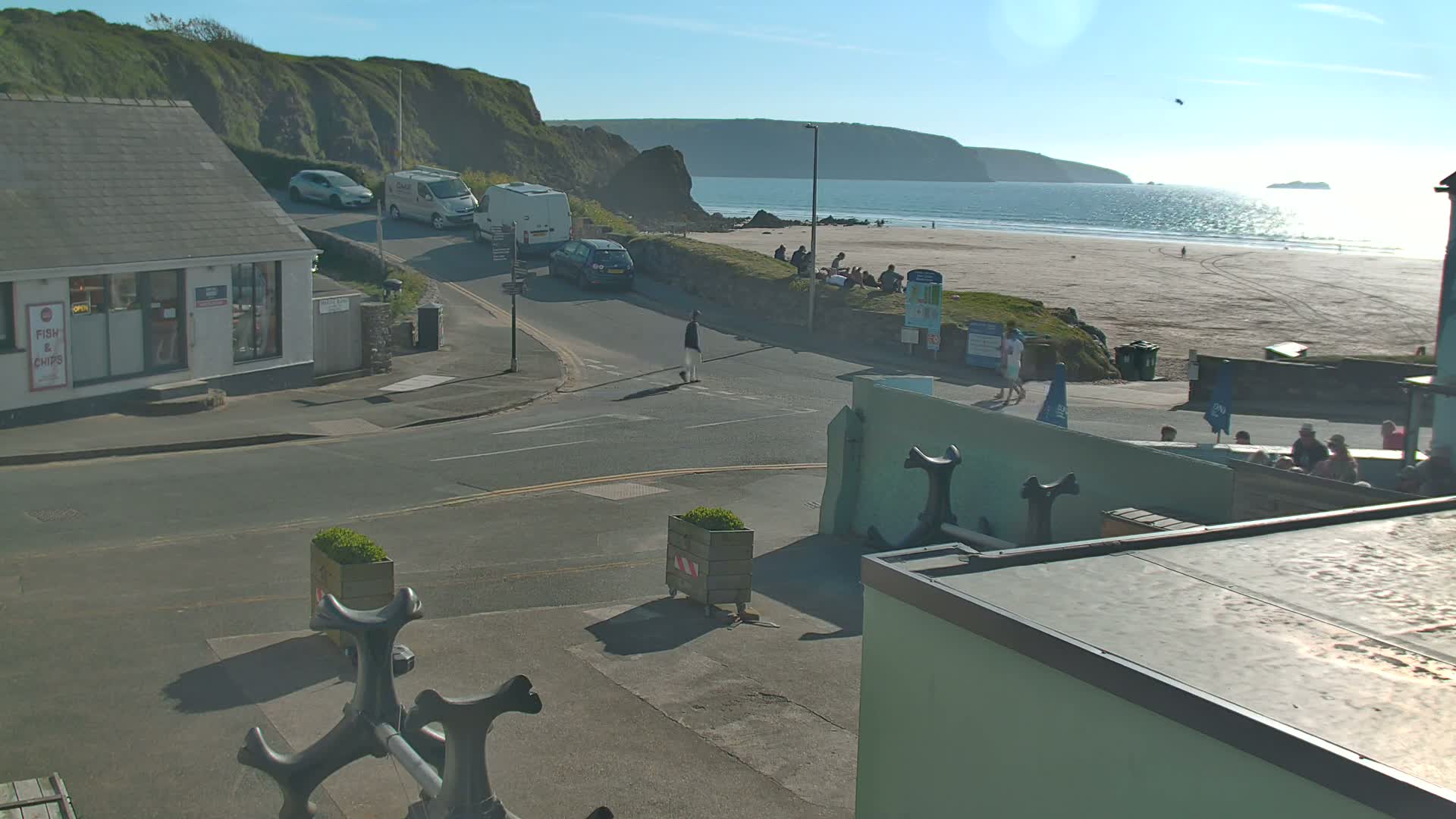 A sunny day reveals a coastal scene with a sandy beach, calm ocean, and a street view of buildings, vehicles, and pedestrians near a road intersection.