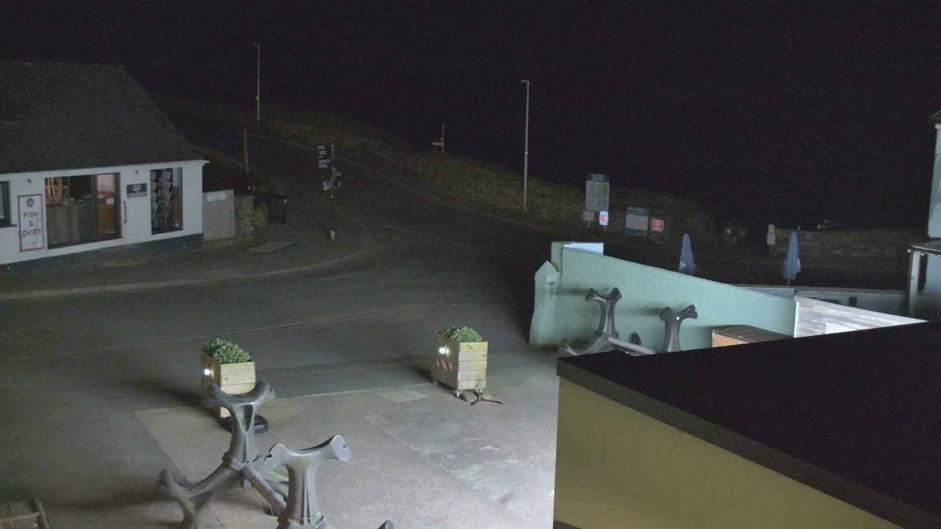 At night, a dimly lit street scene shows a building, several planters, and large anchor sculptures in a mostly empty paved area.