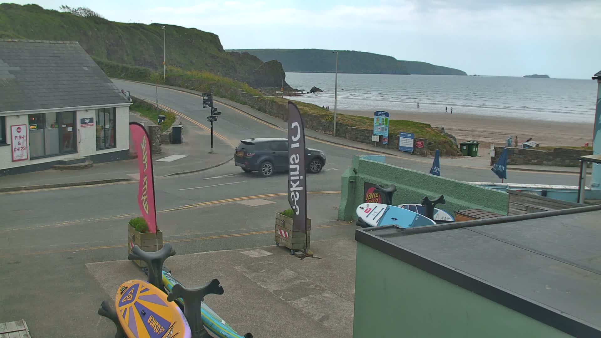 A coastal road scene on an overcast day shows a dark-grey SUV parked near a beach with people visible in the distance, along with surfboards and a fish and chips shop.