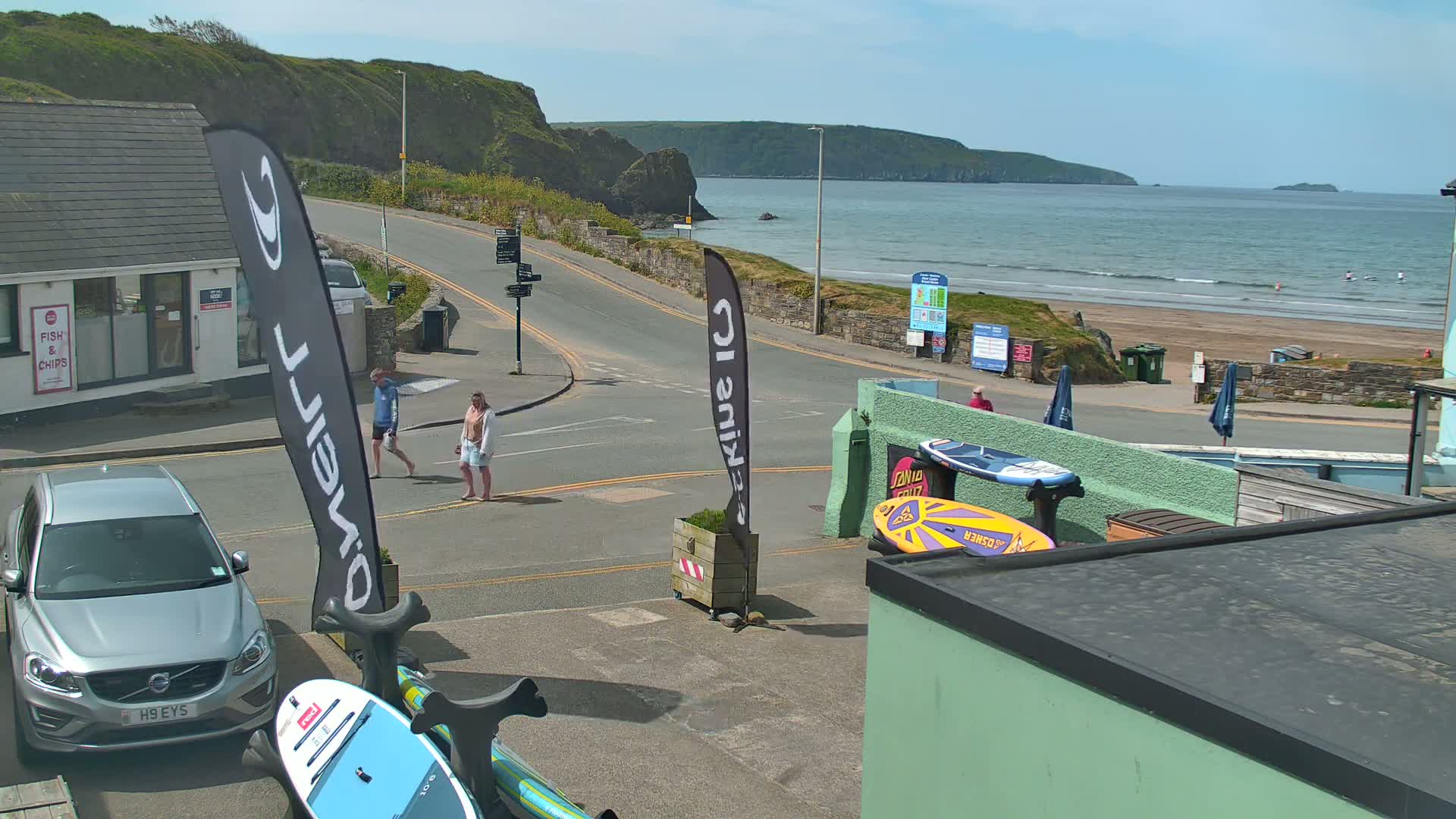 A sunny day at a coastal town shows a road leading to a sandy beach with calm ocean waters, where two people are walking and paddleboards are displayed near a building.