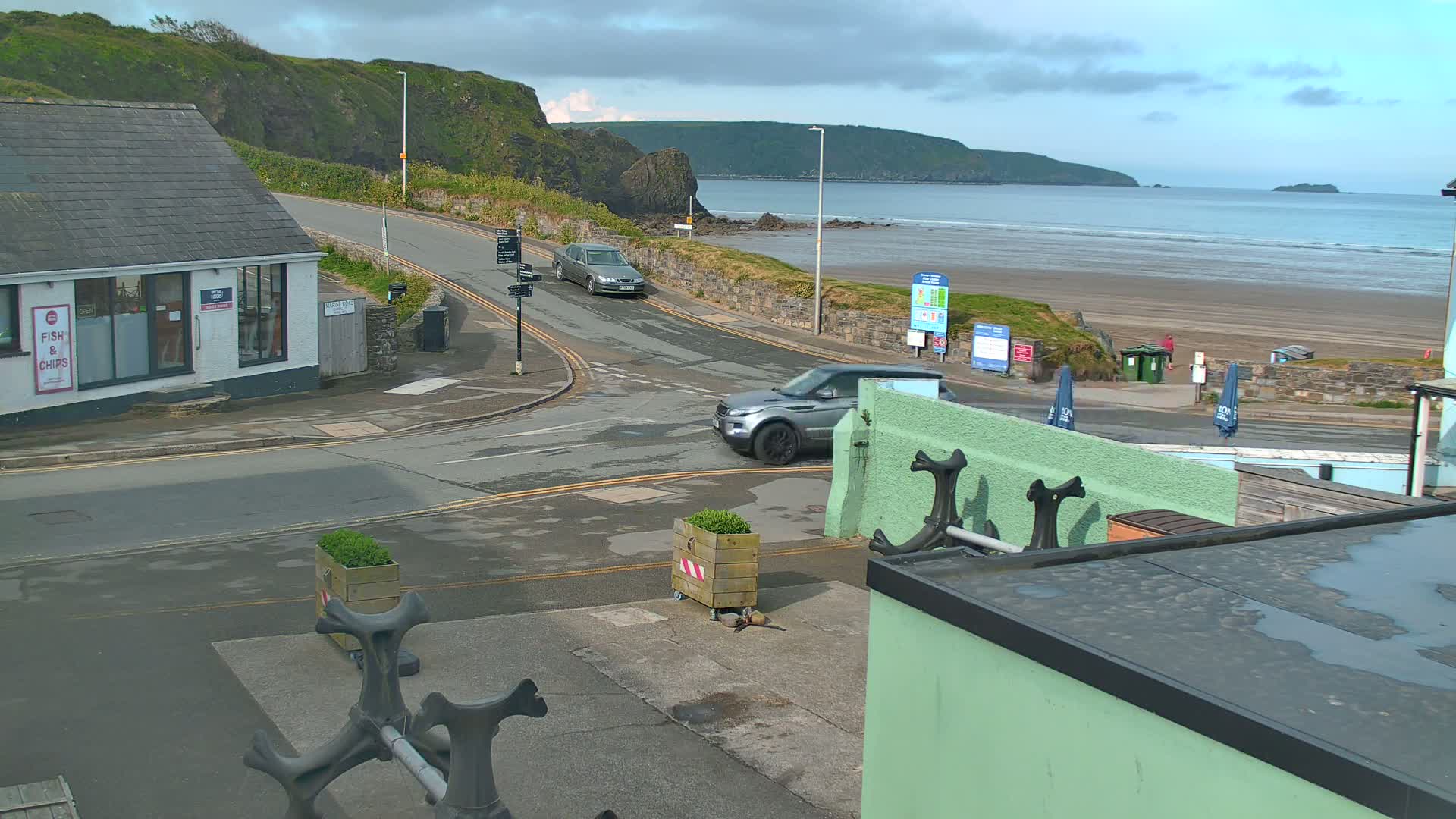 A coastal road scene with two cars, a sandy beach with calm ocean waves, and a building with a fish and chips sign, under a partly cloudy sky.