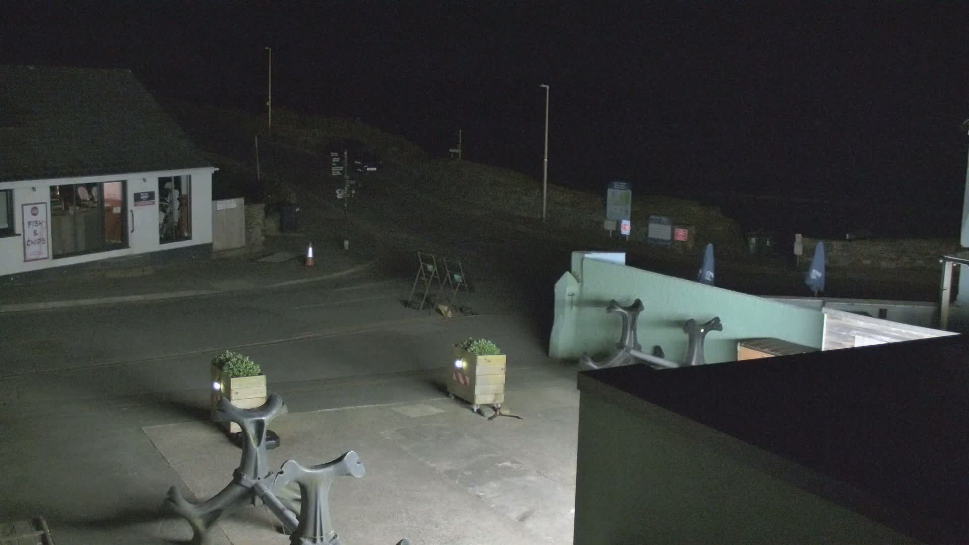 At night, a dimly lit seaside area shows a building, several planters, and outdoor seating near a dark, empty road.