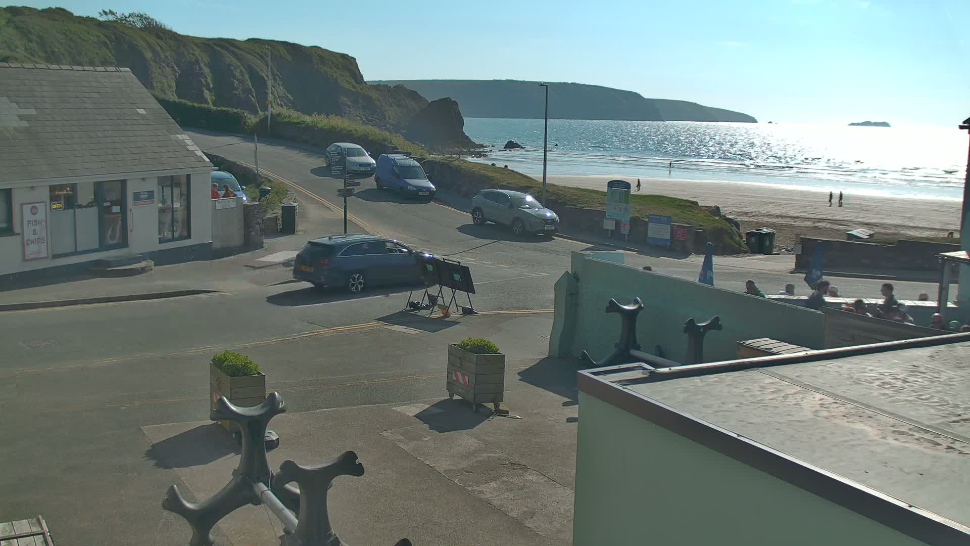 A seaside town scene on a sunny day shows several cars parked and driving near a beach with a few people walking on the sand.