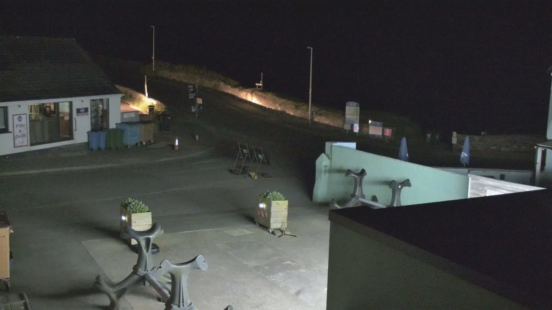 At night, a mostly empty parking lot with a small building and several planters sits under artificial light.