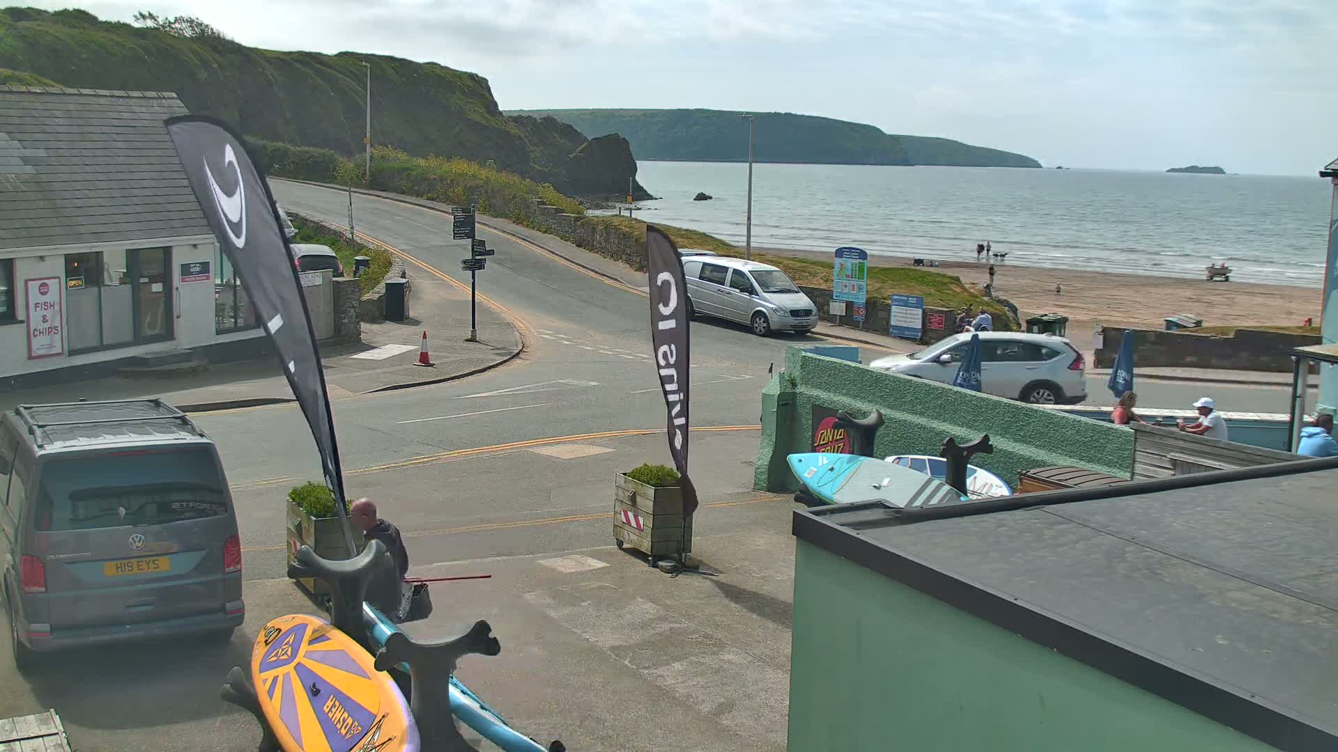 A street scene overlooking a beach on a partly sunny day shows cars parked near a building with surfboards and other equipment, with a body of water and cliffs in the background.