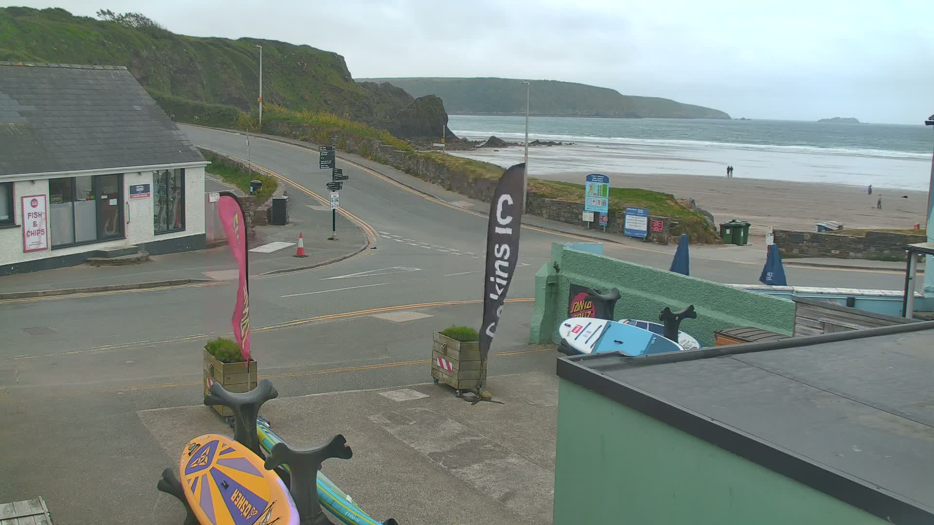 A street scene overlooking a sandy beach with small waves under an overcast sky shows a road curving toward the ocean, with several buildings, and surfboards propped up against a wall.