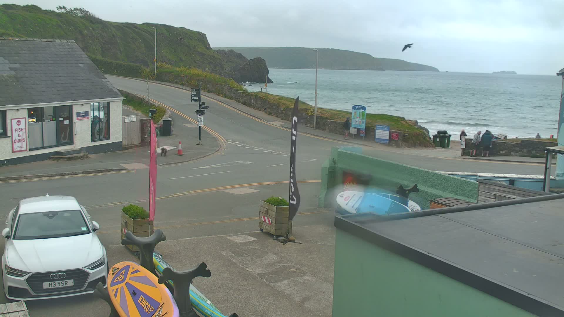 A coastal road curves toward a beach with a few people visible, under an overcast sky; a white car is parked near surfboards.