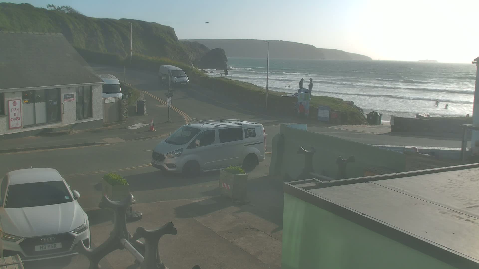 A street scene overlooking a beach with moderate waves under a hazy, sunny sky, features several vehicles including a white van and a white car, and a few people are visible near the shore.