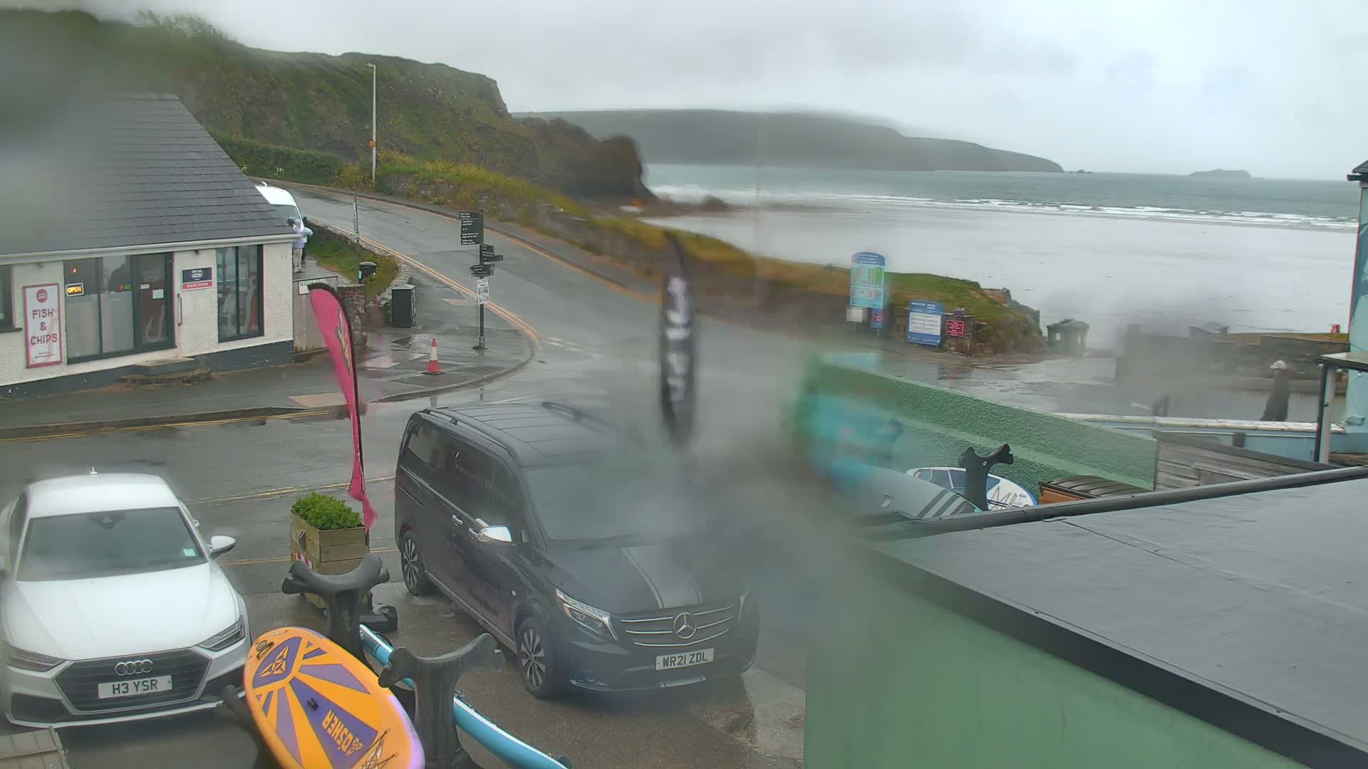 A rainy coastal scene shows a street corner with parked cars, a beach in the distance, and a building with a "Fish & Chips" sign.