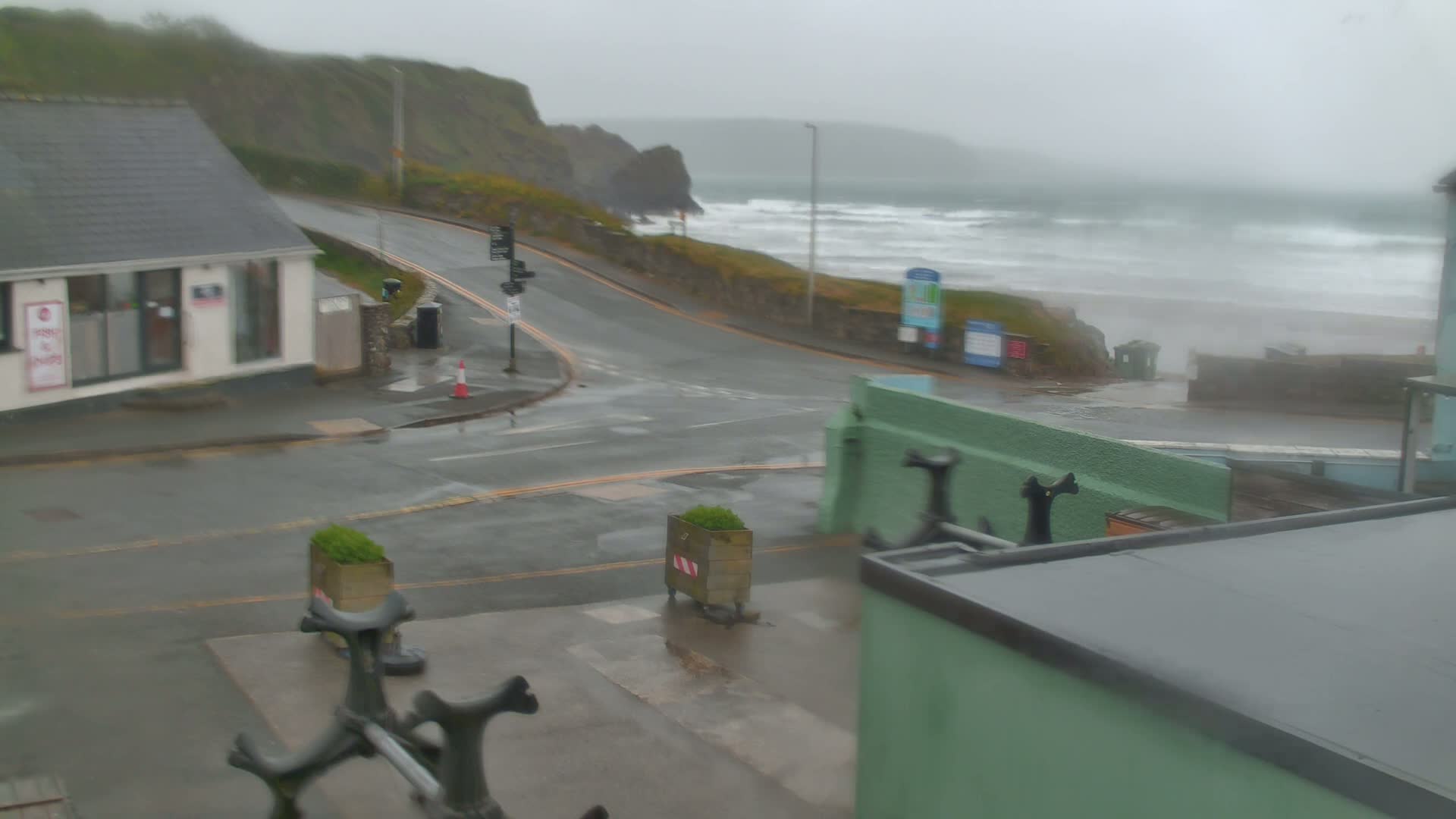 A wet coastal road curves around a building toward a beach with heavy waves under a gray, overcast sky.
