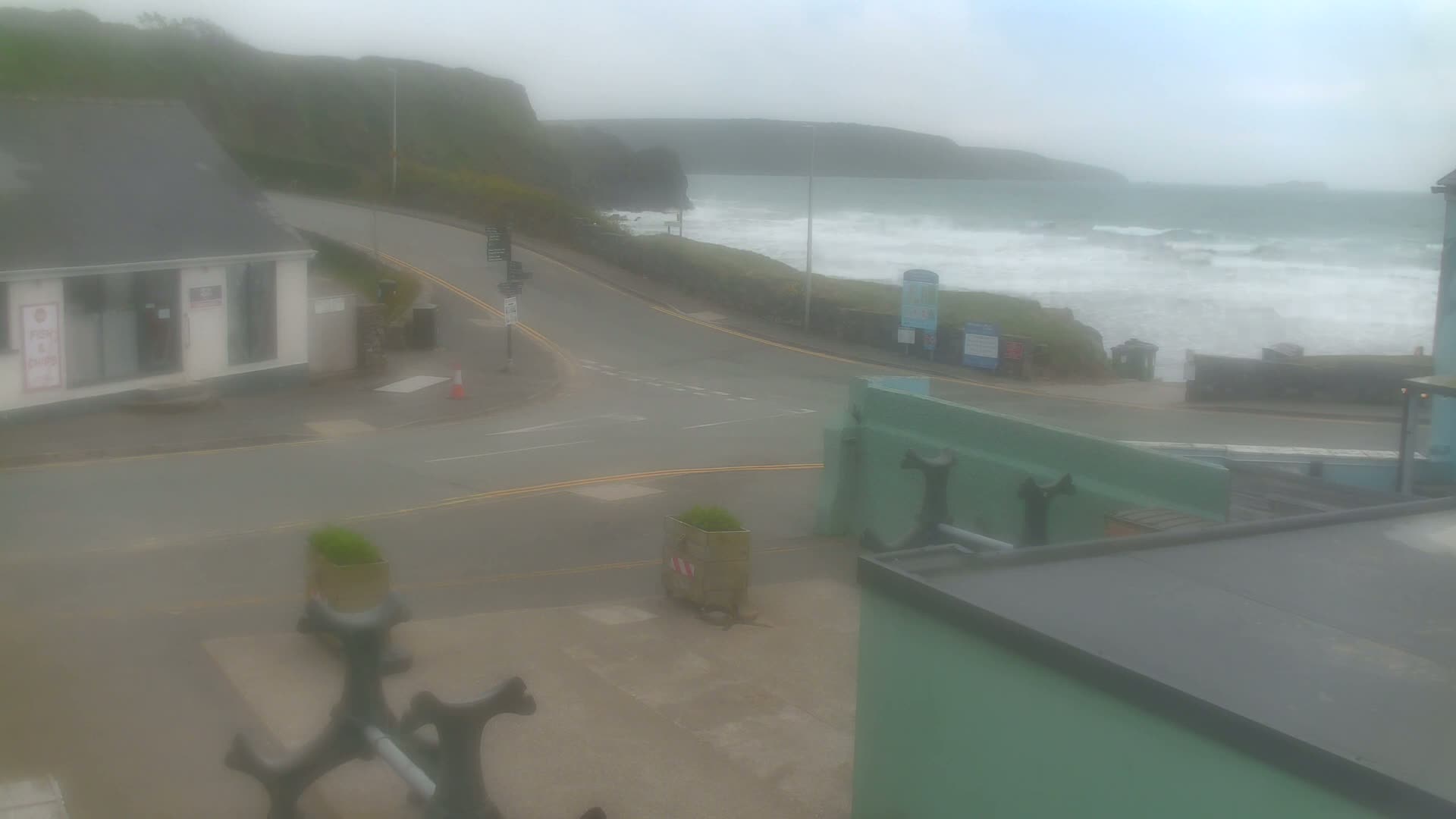 A foggy coastal road intersects near a building overlooking a choppy sea with waves crashing against the shore.