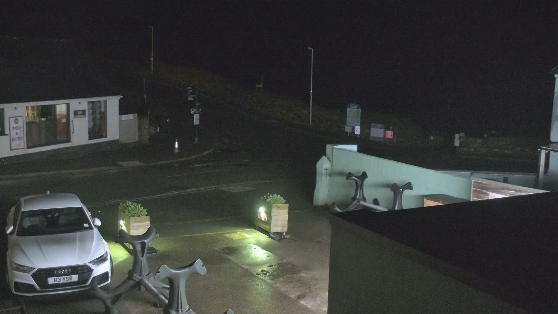 A white car is parked in a dark, wet parking lot at night, near a building and several decorative anchors.