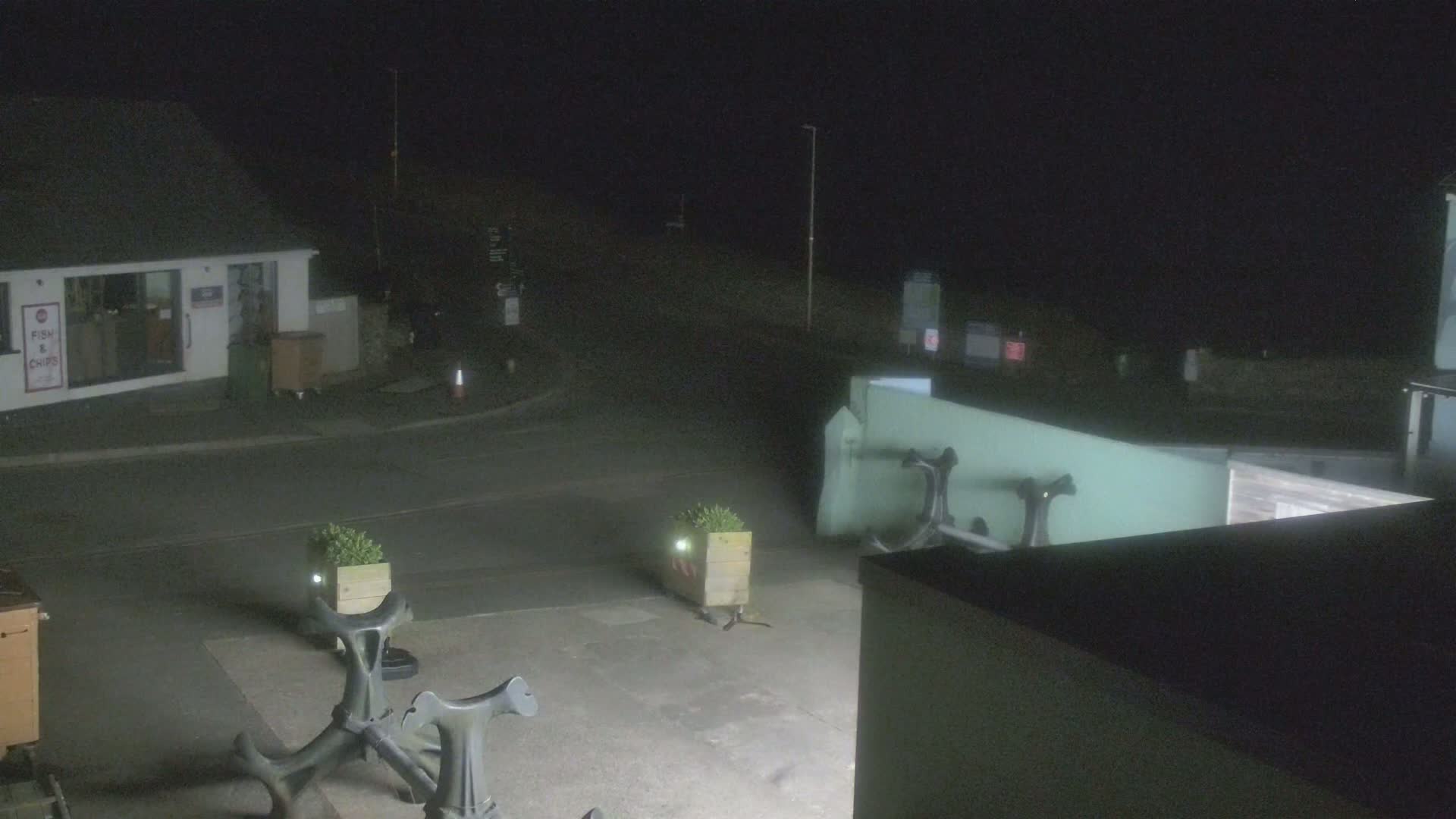 At night, a mostly empty street scene shows a small building, some planters, and outdoor seating near what appears to be a harbor.