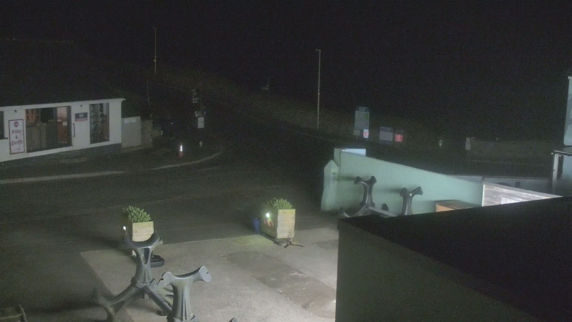 A nighttime, low-light view of an empty street corner with a building and several dark, metal objects that resemble anchors.