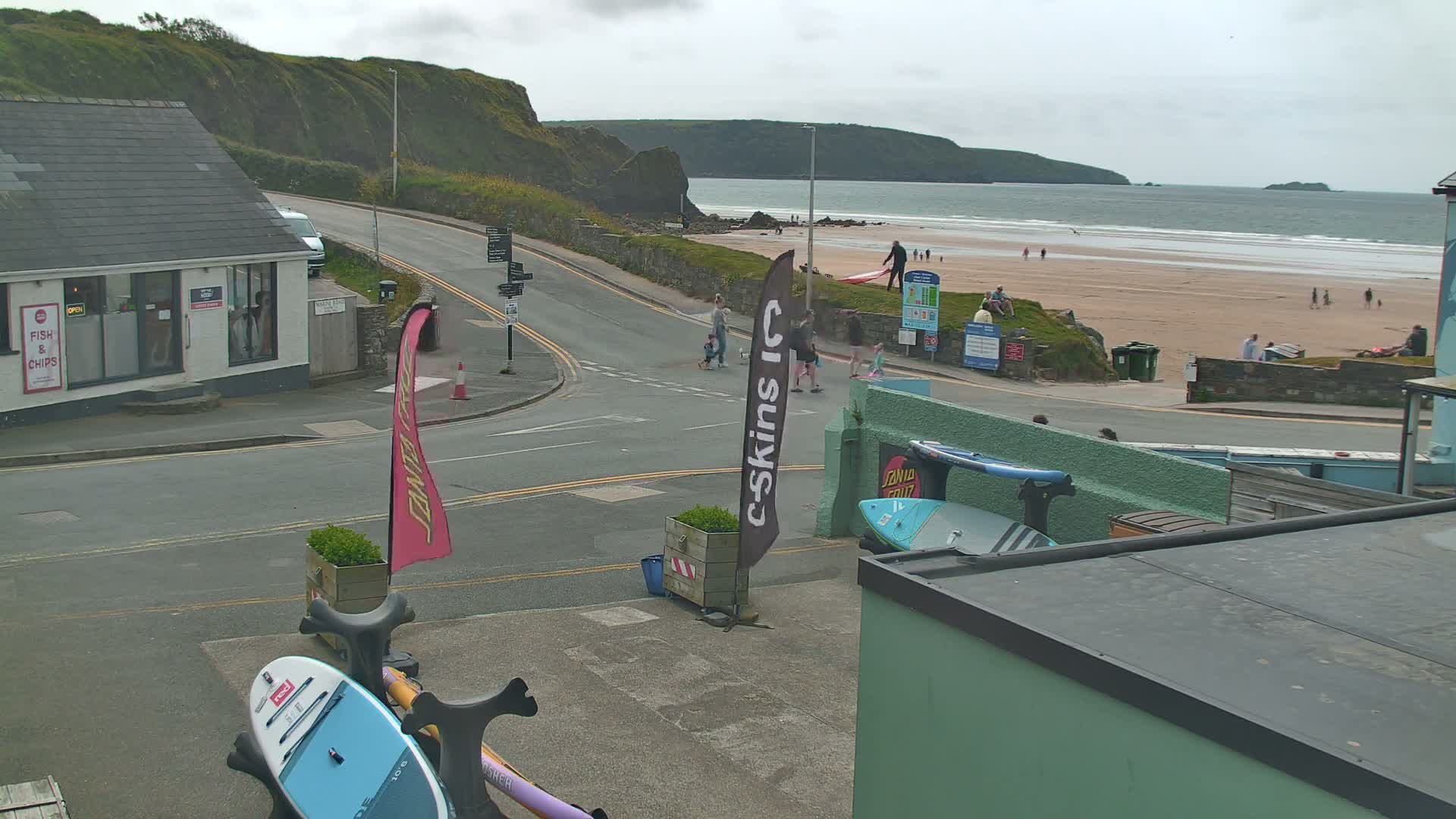 A street scene overlooks a sandy beach with calm ocean waves under an overcast sky; several people are visible on the beach and street.