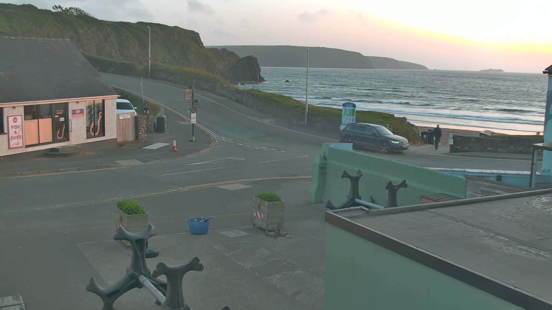 A coastal road scene at sunset shows a car parked near a beach with gentle waves, a small building, and some landscaping.