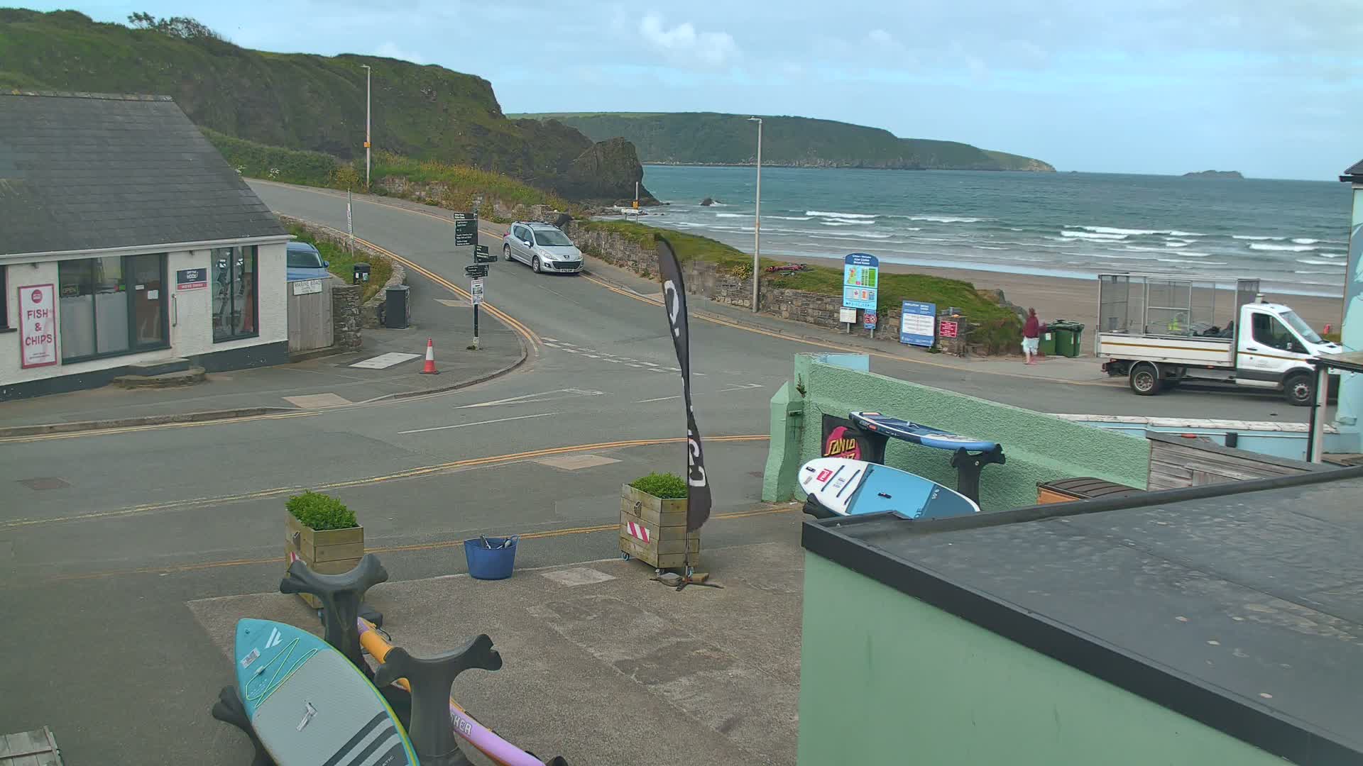 A seaside scene on a cloudy day shows a road curving toward a beach with moderate waves, a car parked nearby, and surfboards resting near a building.