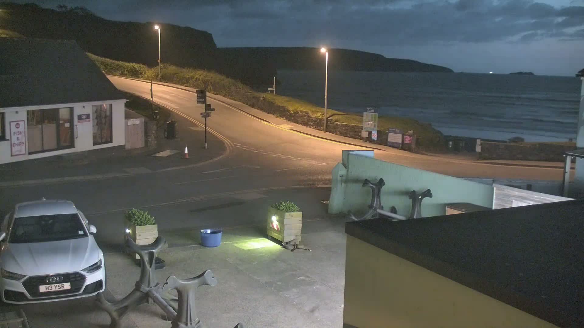 A white car is parked in a seaside town at night, with a dark, wavy ocean visible in the background under a cloudy sky.
