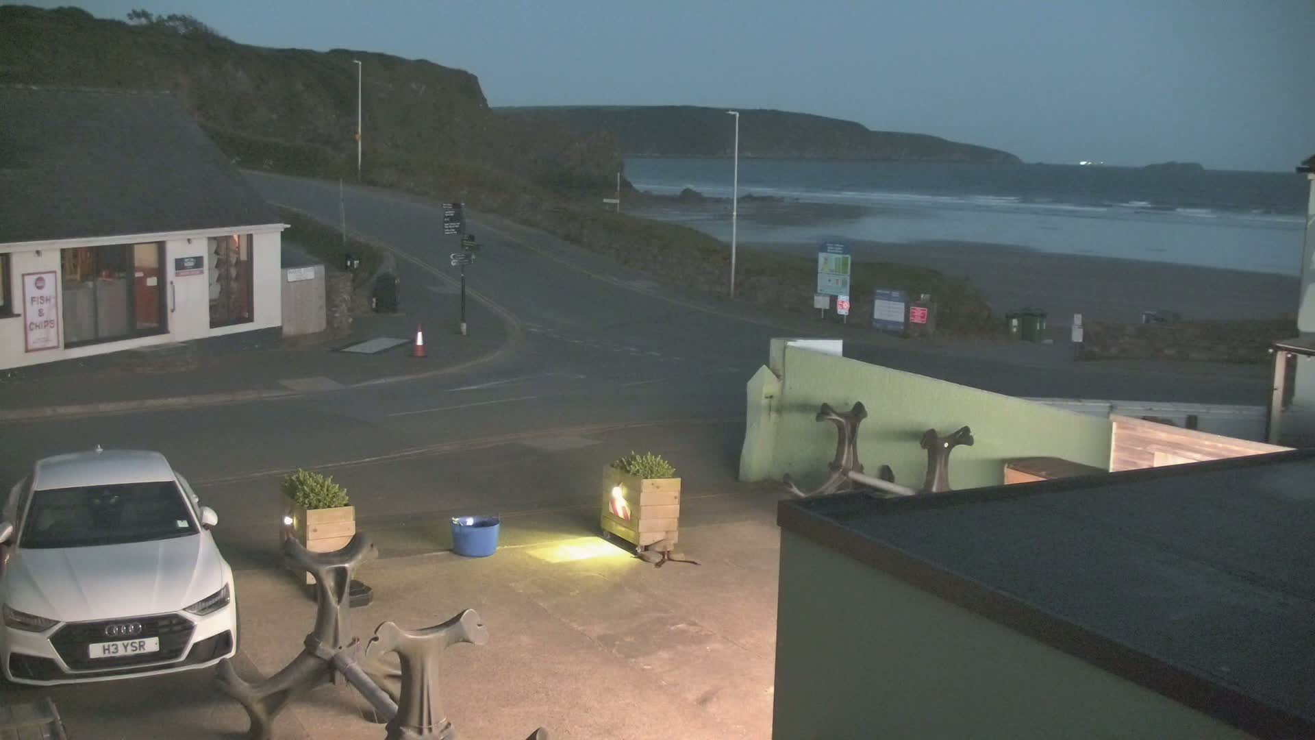 A white car is parked near a coastal road at dusk, overlooking a beach with gentle waves under a dark sky.