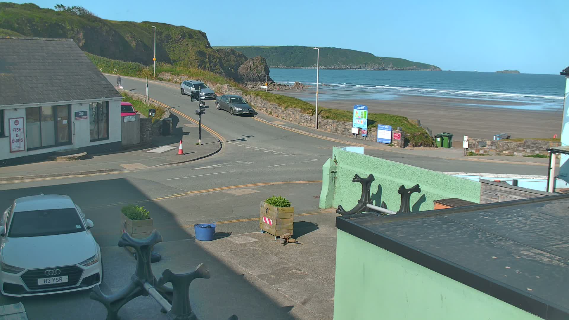 A sunny day at a coastal town shows a sandy beach with waves, several cars on a road leading to it, and buildings, including a small shop.