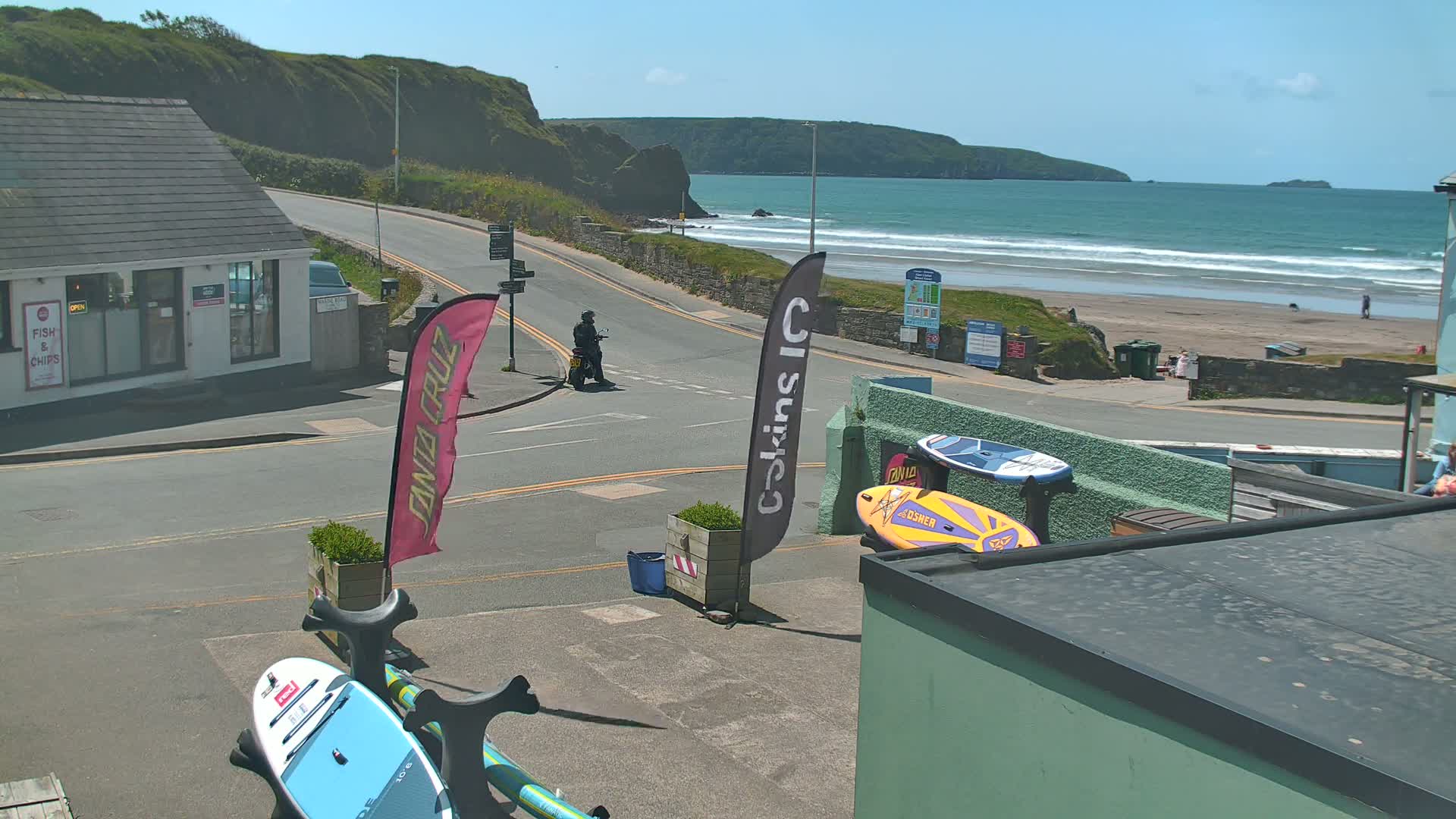 A seaside town scene on a sunny day shows a paved road curving toward a sandy beach with waves rolling in, alongside various shops and surfboards.