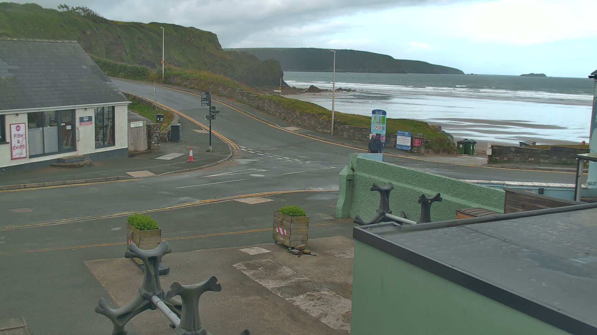 A coastal road intersects near a fish and chips shop, overlooking a beach with gently rolling waves under a cloudy sky.