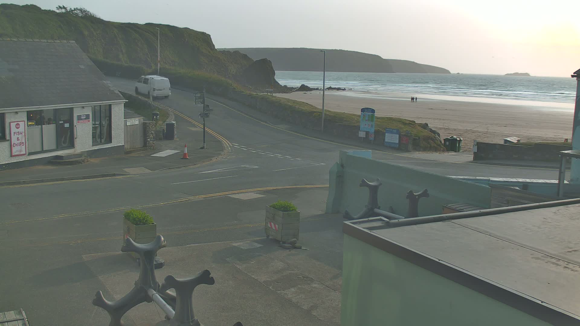 A road curves toward a sandy beach with small waves under a hazy sky, next to a building with a fish and chips shop.