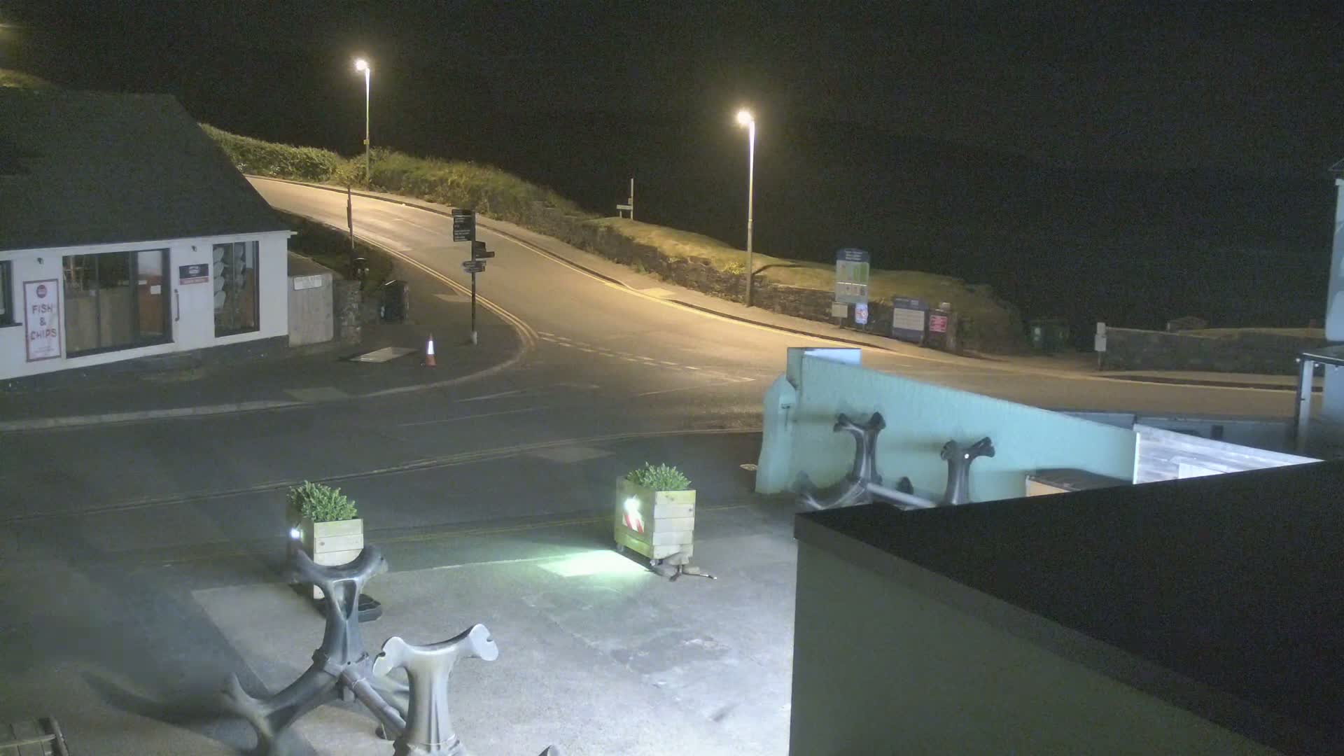 At night, a street curves along a cliffside, past a small building and a plaza with large anchor sculptures;  the street is dimly lit by streetlights.