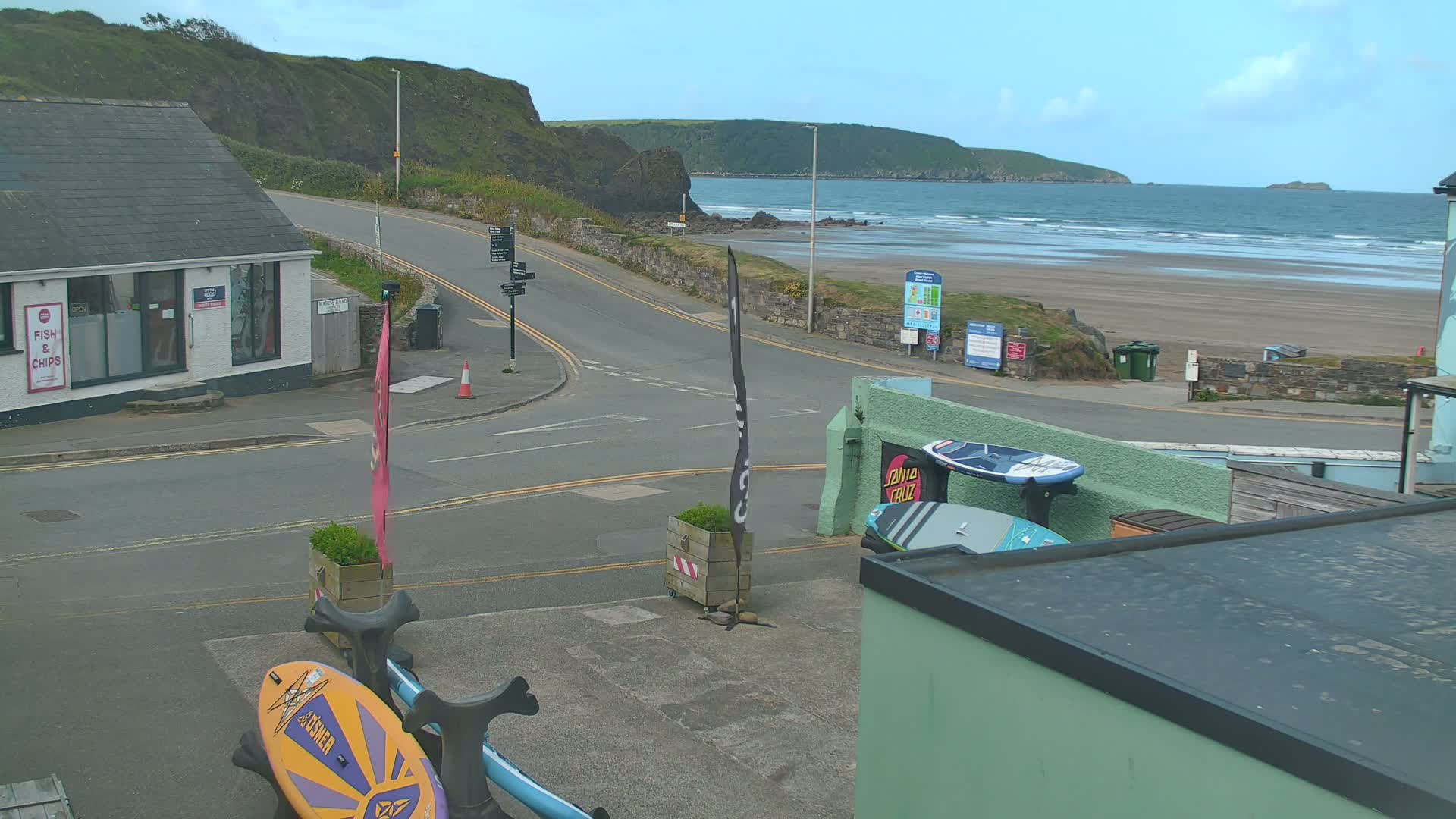 A coastal town scene on a partly cloudy day shows a road curving toward a sandy beach with gentle waves,  surfboards are visible near a building.
