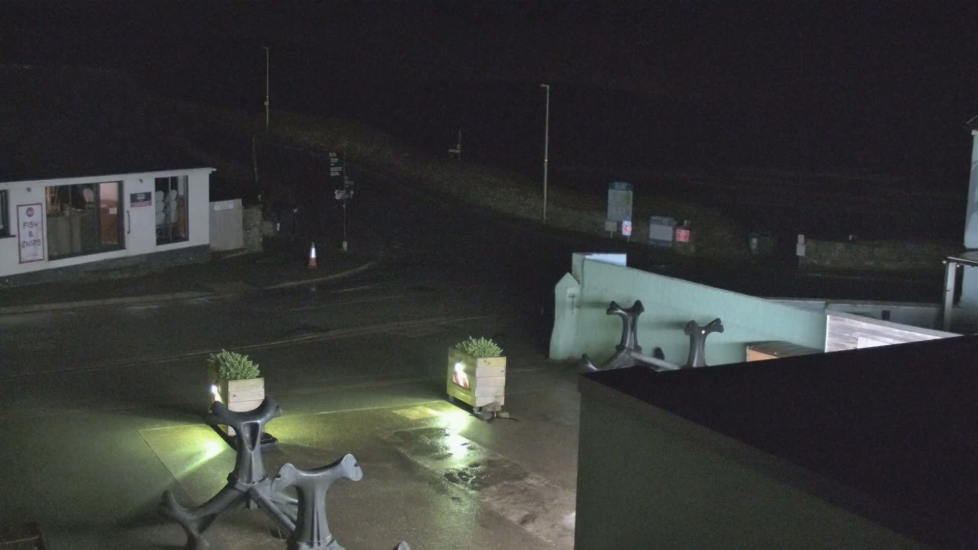 At night, a wet, dark parking lot is illuminated by lights near two decorative anchors and potted plants, adjacent to a building and a low wall.
