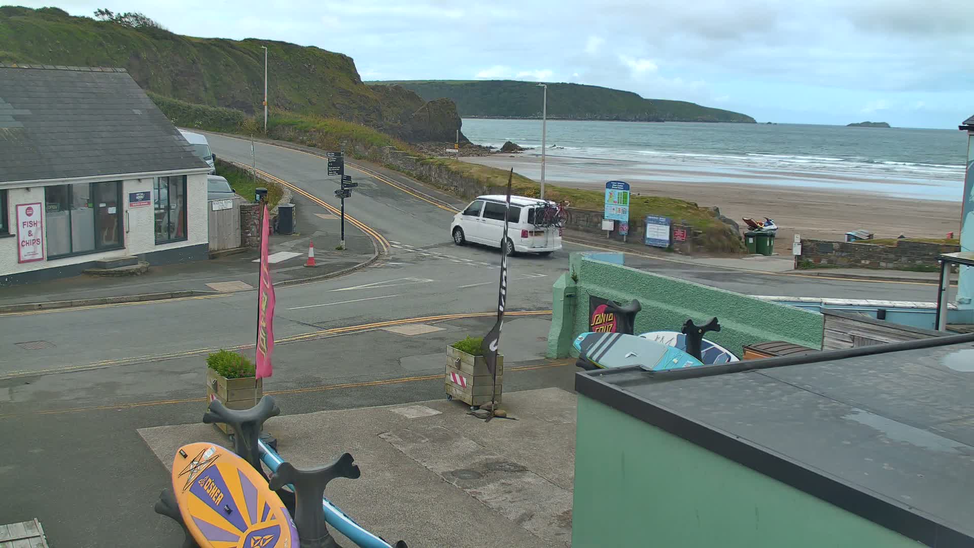 A white van drives past a beach with small waves under an overcast sky, near buildings including a fish and chips shop and surf shops with surfboards displayed.