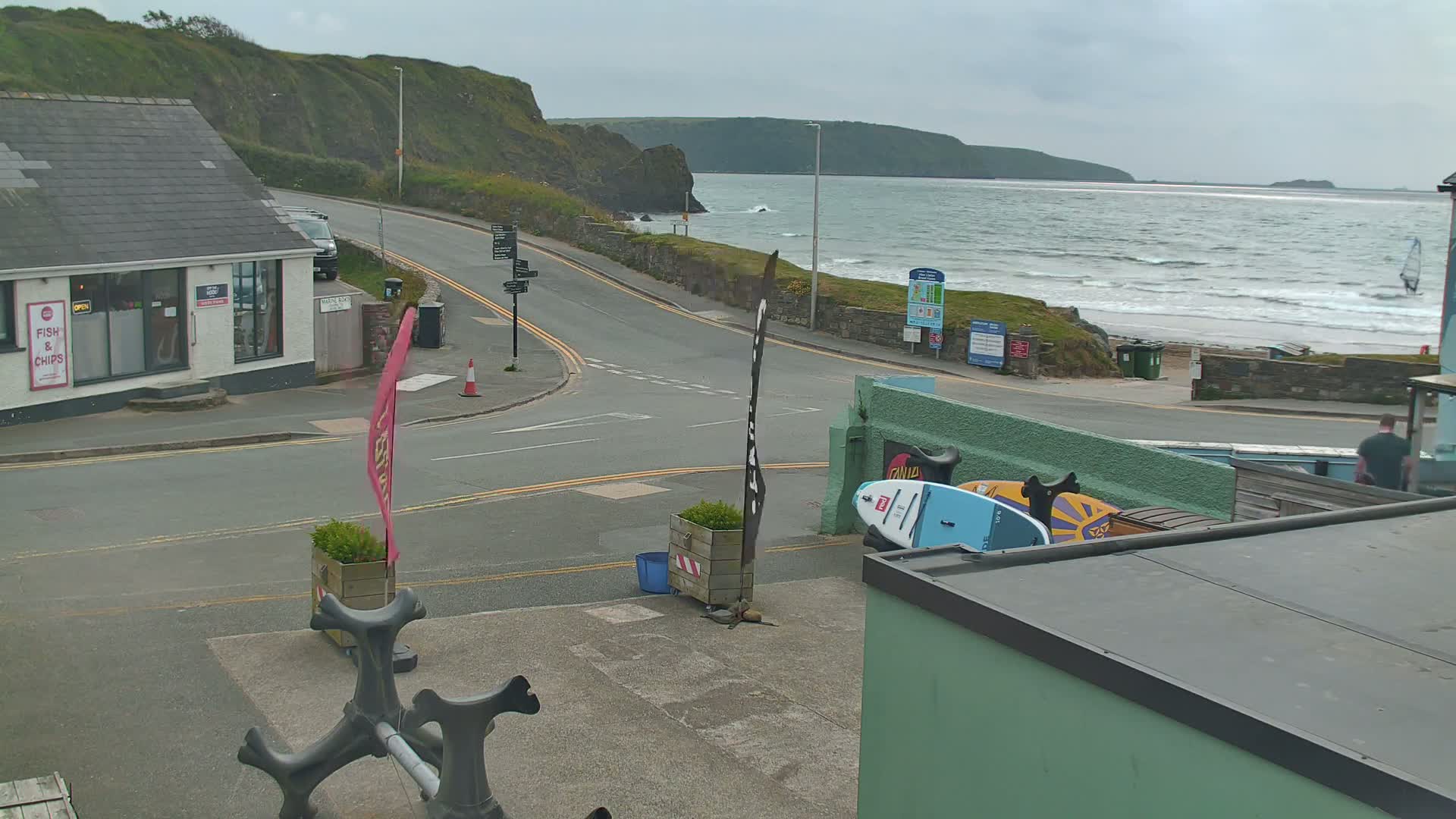 A coastal town scene shows a road intersecting near a fish and chips shop, with a wavy ocean and a beach visible in the background under an overcast sky.