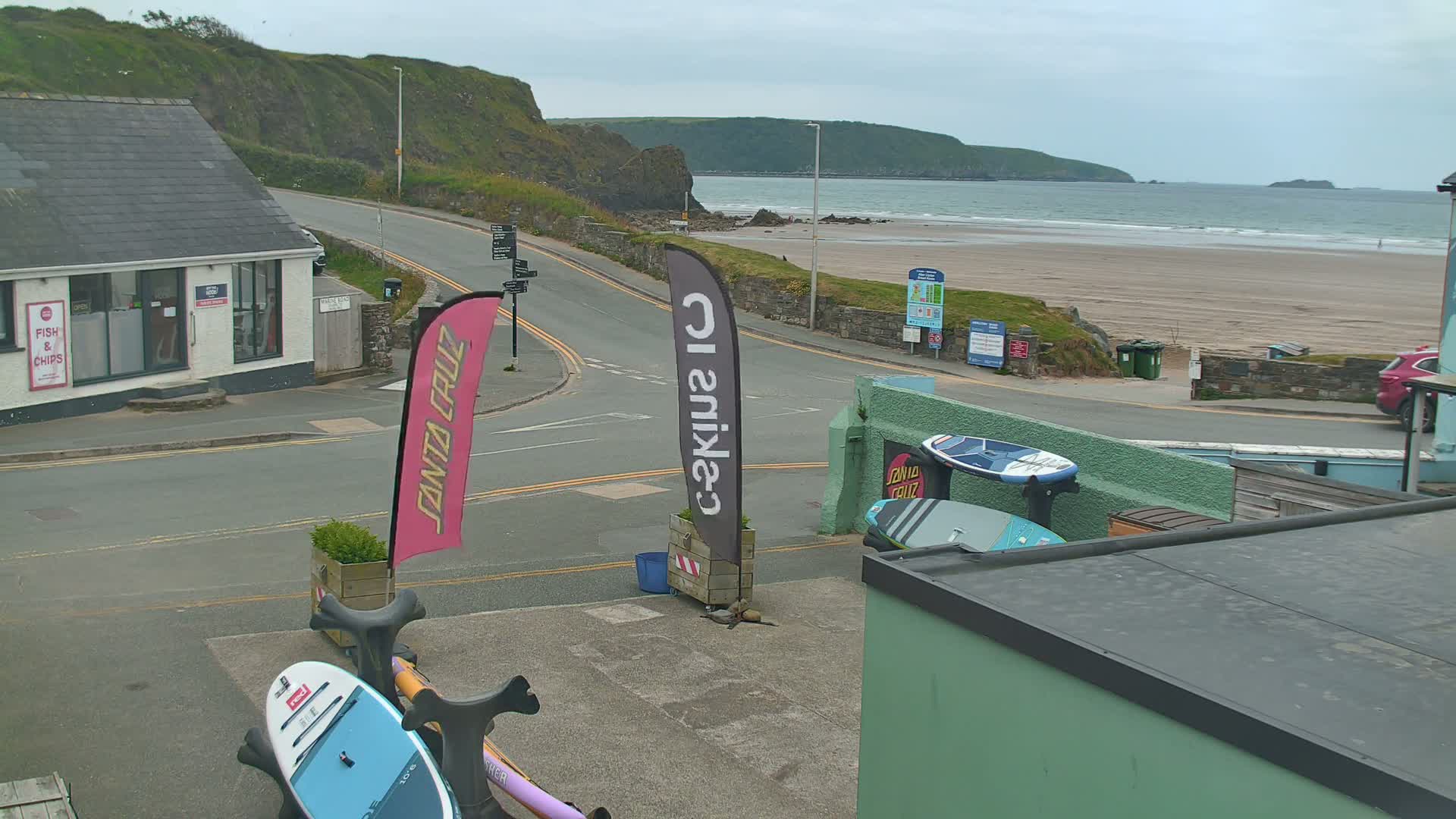 A street scene overlooking a sandy beach on an overcast day shows a road curving toward the water, several paddleboards, and a building with a “Fish & Chips” sign.