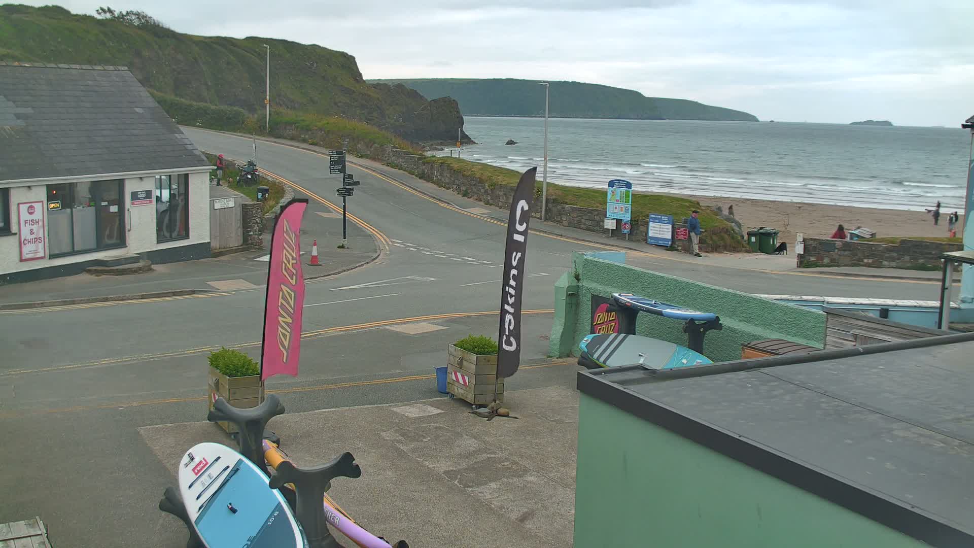 A seaside town scene on an overcast day shows a road curving toward a beach with people and surfboards visible.