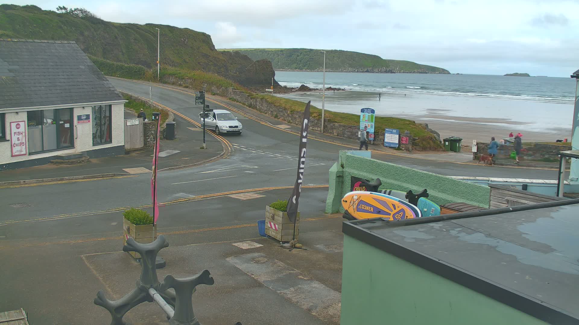 A street scene overlooking a beach on an overcast day shows a car at an intersection near a fish and chips shop, with people walking on the beach in the distance.