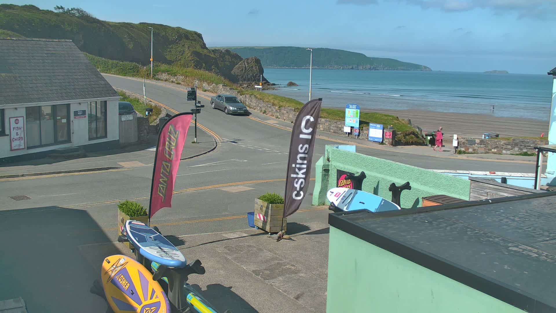 A sunny day at a coastal town shows a sandy beach with calm waves, a car driving on a road near a building, and surfboards leaning against a structure.