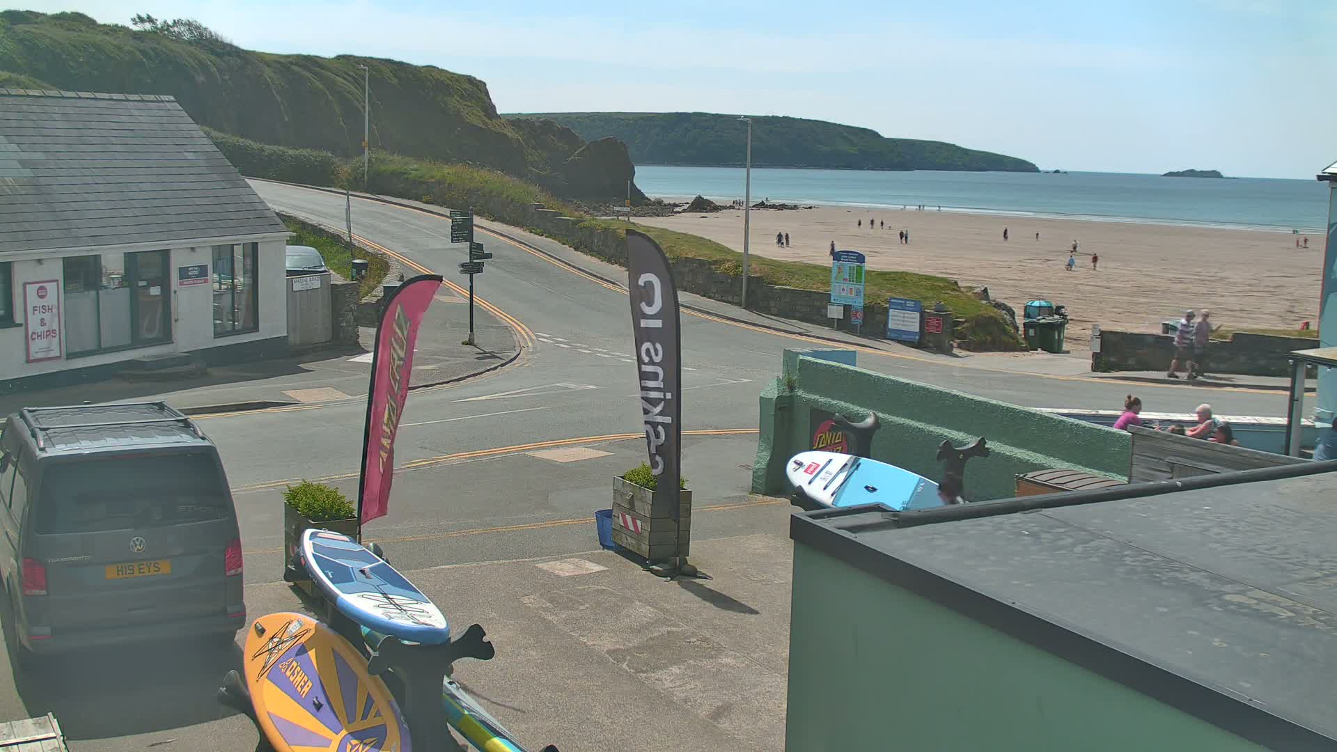 A sunny day at a beach viewed from a street corner, showing a sandy beach with several people, a road with cars, and paddleboards leaning against a building.