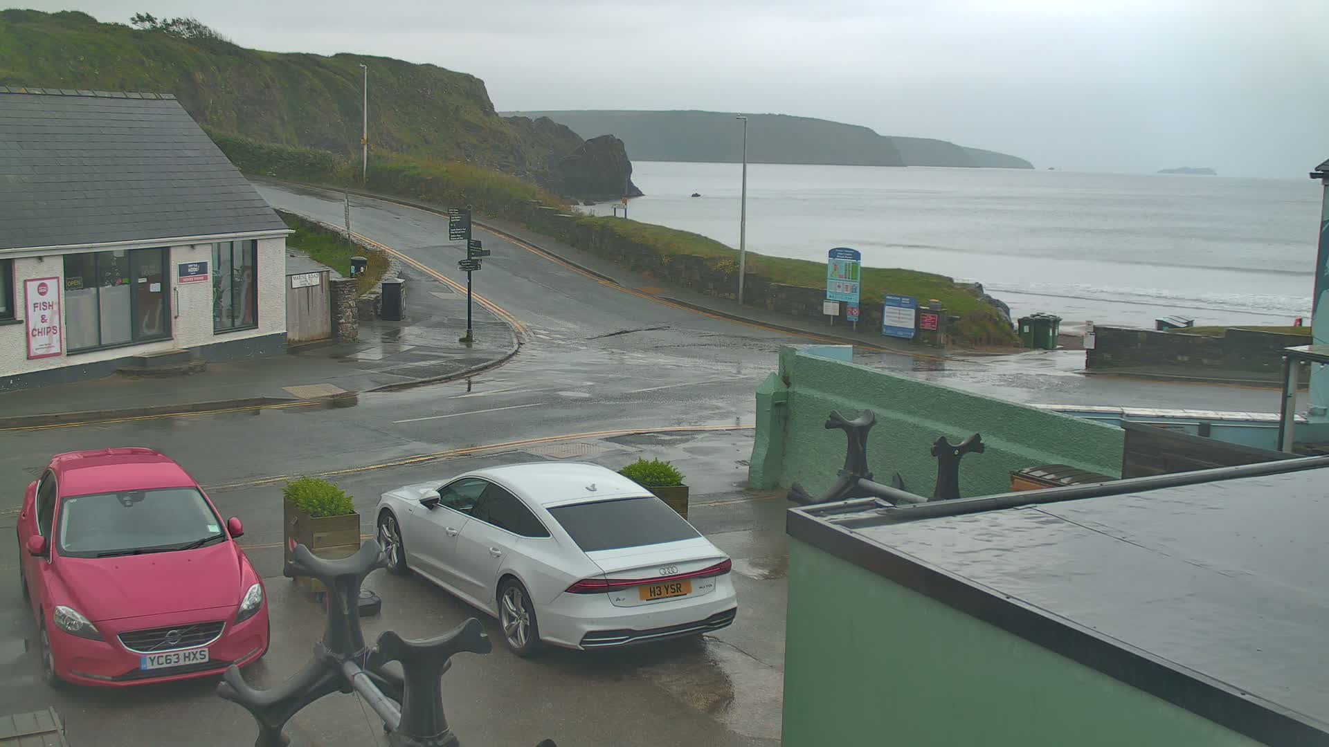 A wet, overcast day at a coastal town shows a street intersection with two cars parked near a building, overlooking a calm sea with small waves and cliffs in the distance.