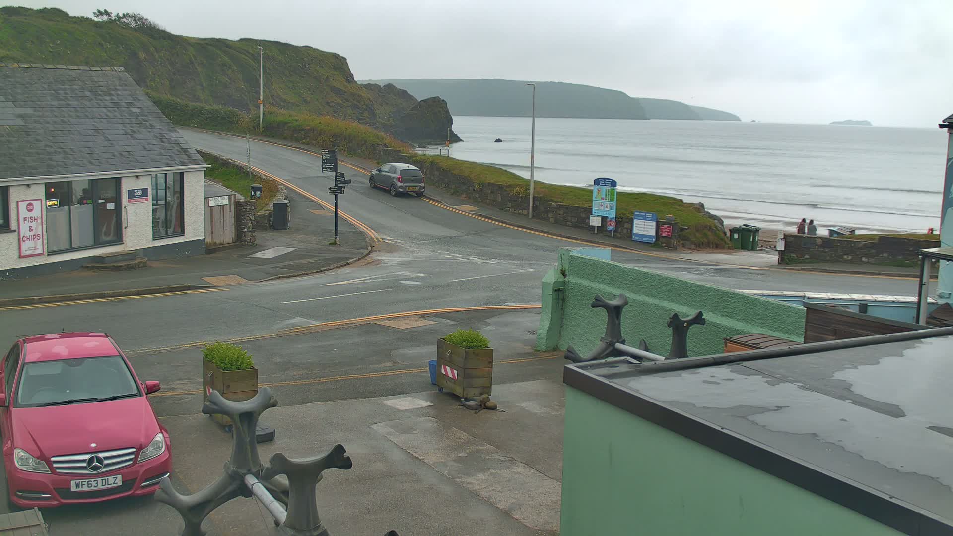 A coastal road intersection with a pink car parked nearby, overlooking a gray, overcast beach with small waves.