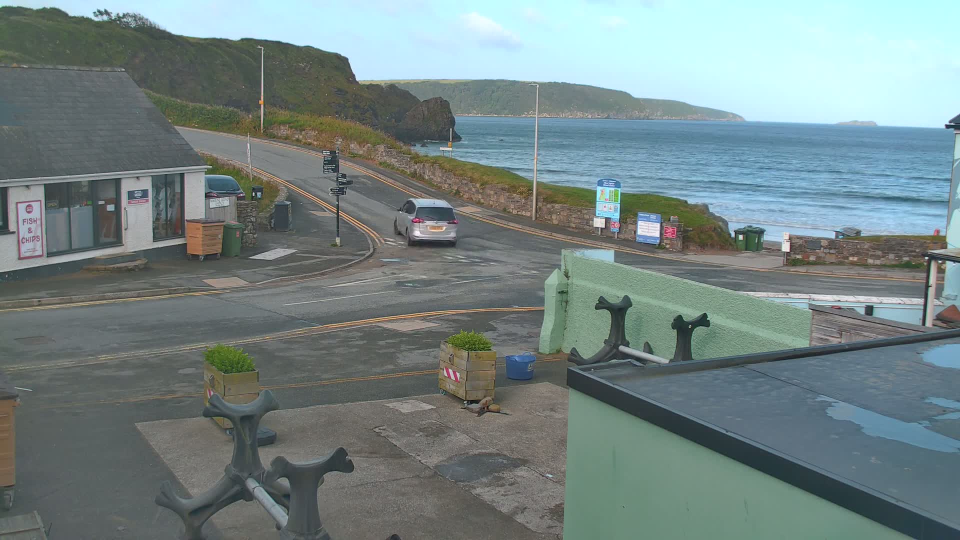 A silver car drives on a road overlooking a beach with gently rolling waves under a clear sky.