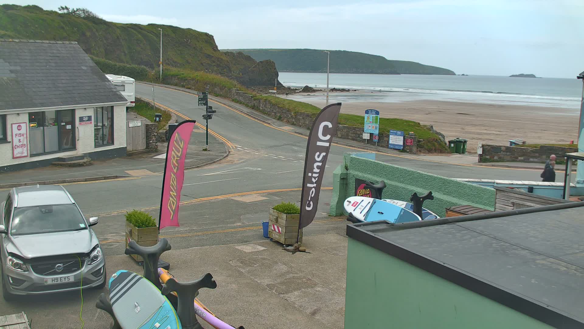 A street scene overlooking a sandy beach with gentle waves under an overcast sky shows a car parked near a shop and surfboards leaning against a building.