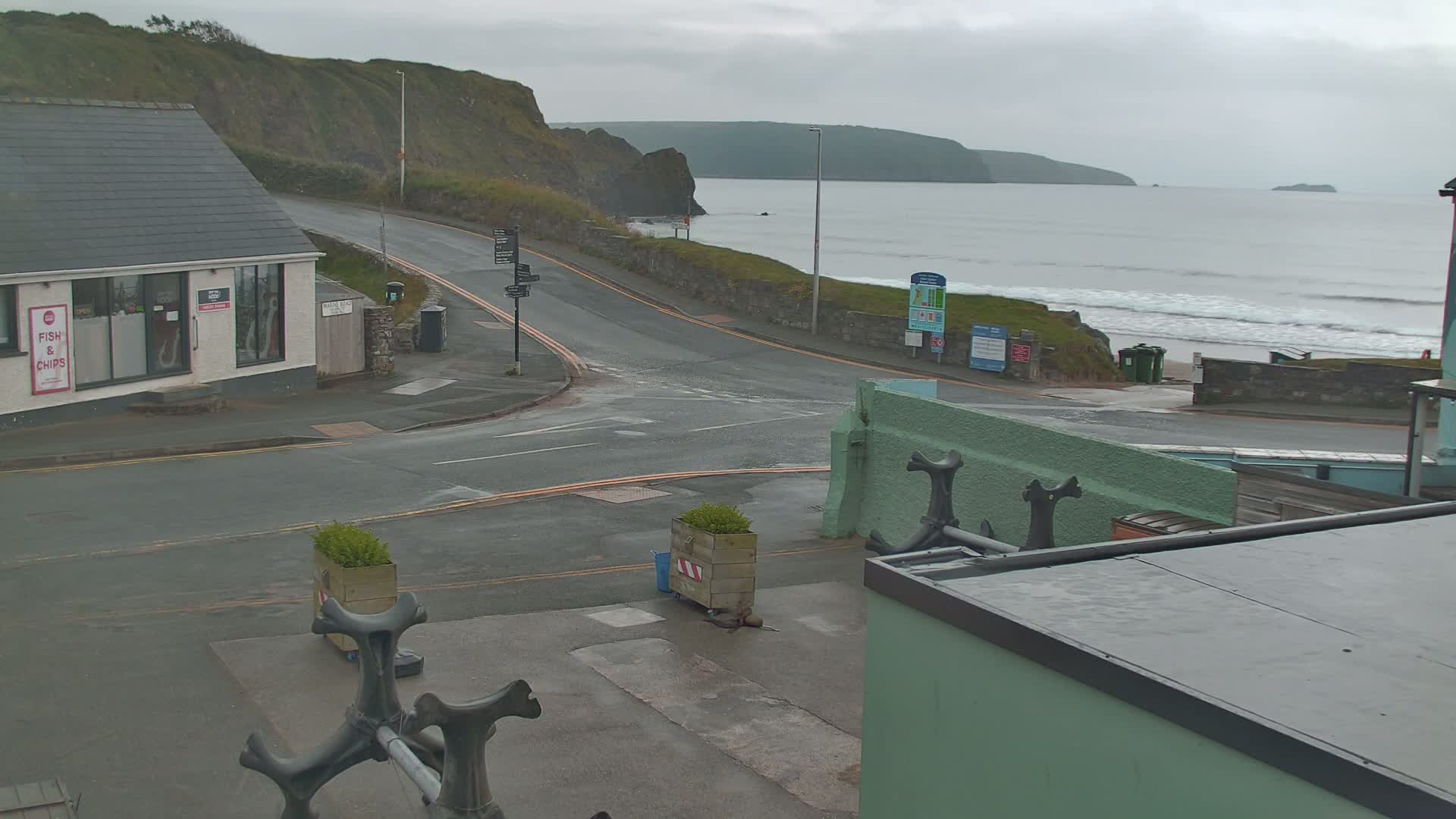 A seaside road with a fish and chips shop and a view of a grey, overcast ocean with small waves crashing on a beach.
