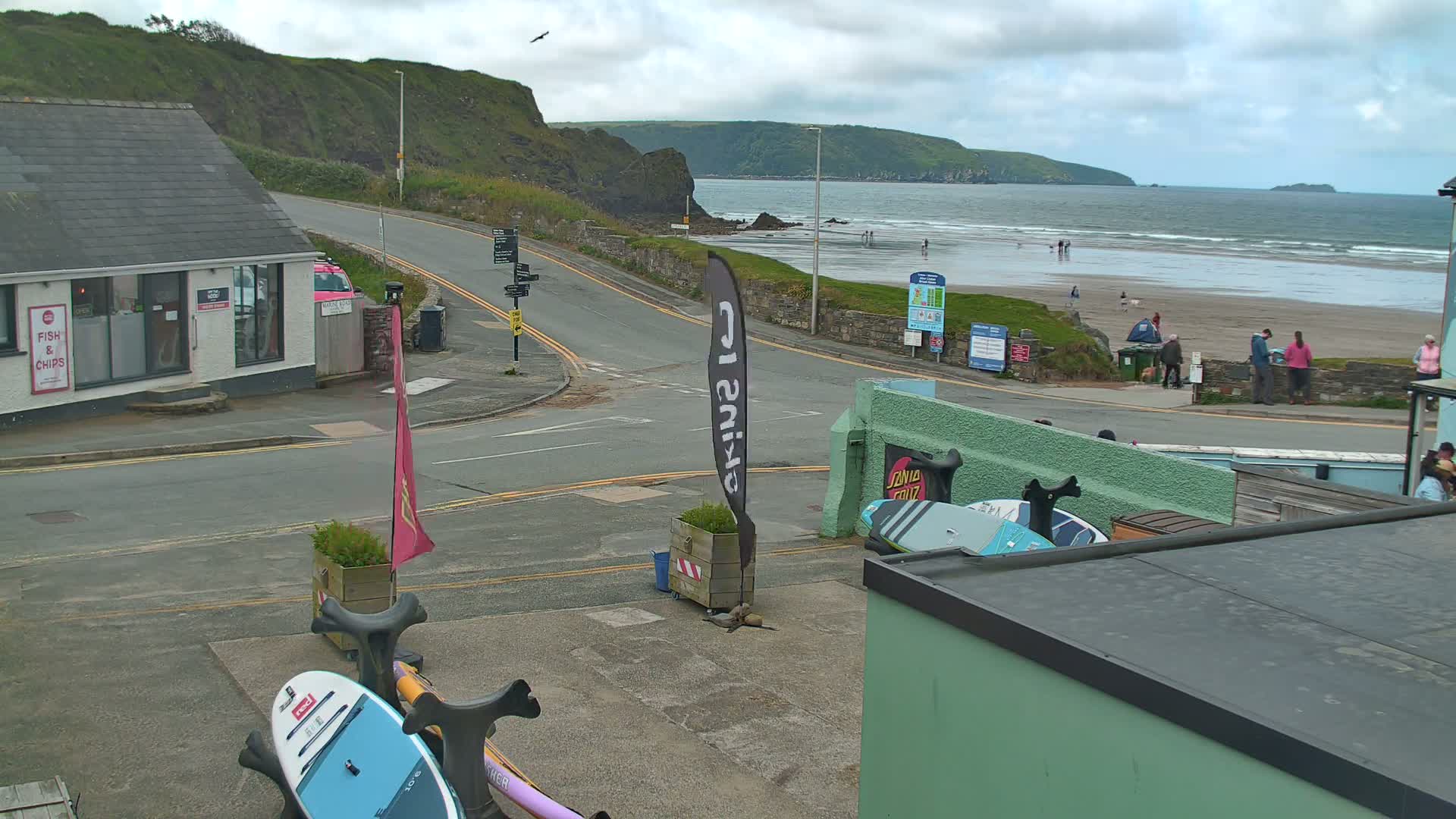 A street scene overlooking a beach with overcast skies shows a road curving toward the ocean, where several people are standing on the sand, and paddleboards are leaning against a building.