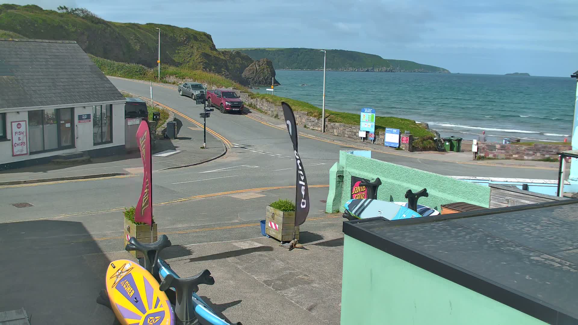 A sunny day overlooking a coastal road with parked cars, a beach with light waves, and a rocky hillside.