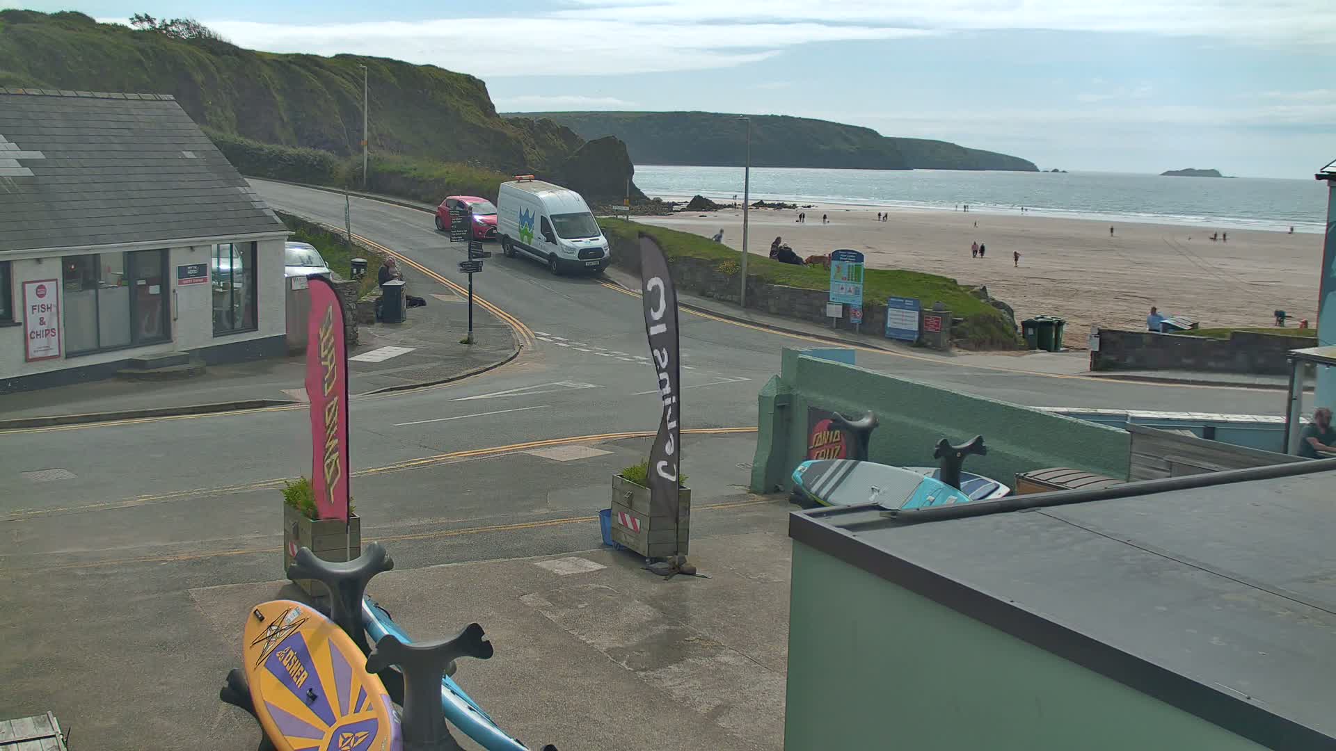 A seaside town scene on a partly cloudy day shows a road curving toward a sandy beach with people on it, backed by a rocky coastline.