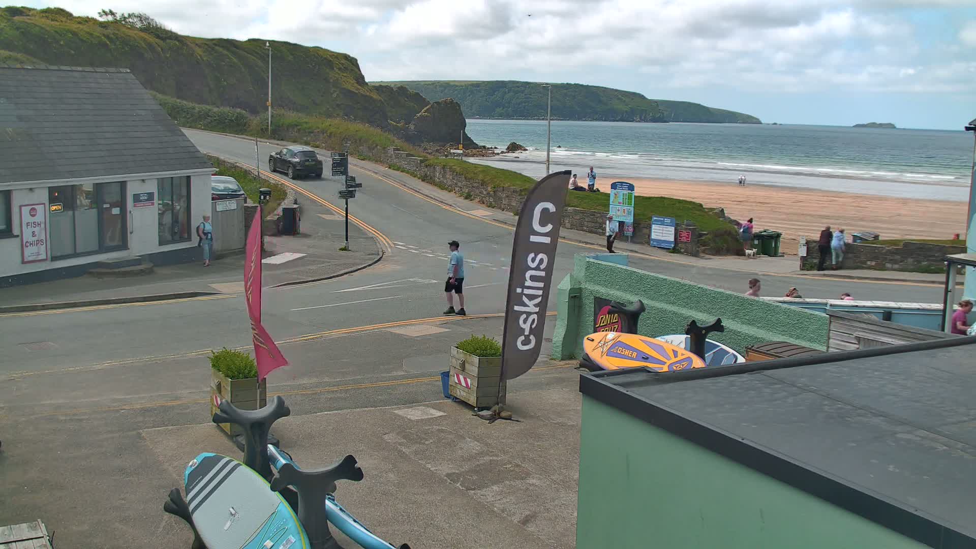 A view of a coastal road overlooking a sandy beach with calm ocean waves under a partly cloudy sky.